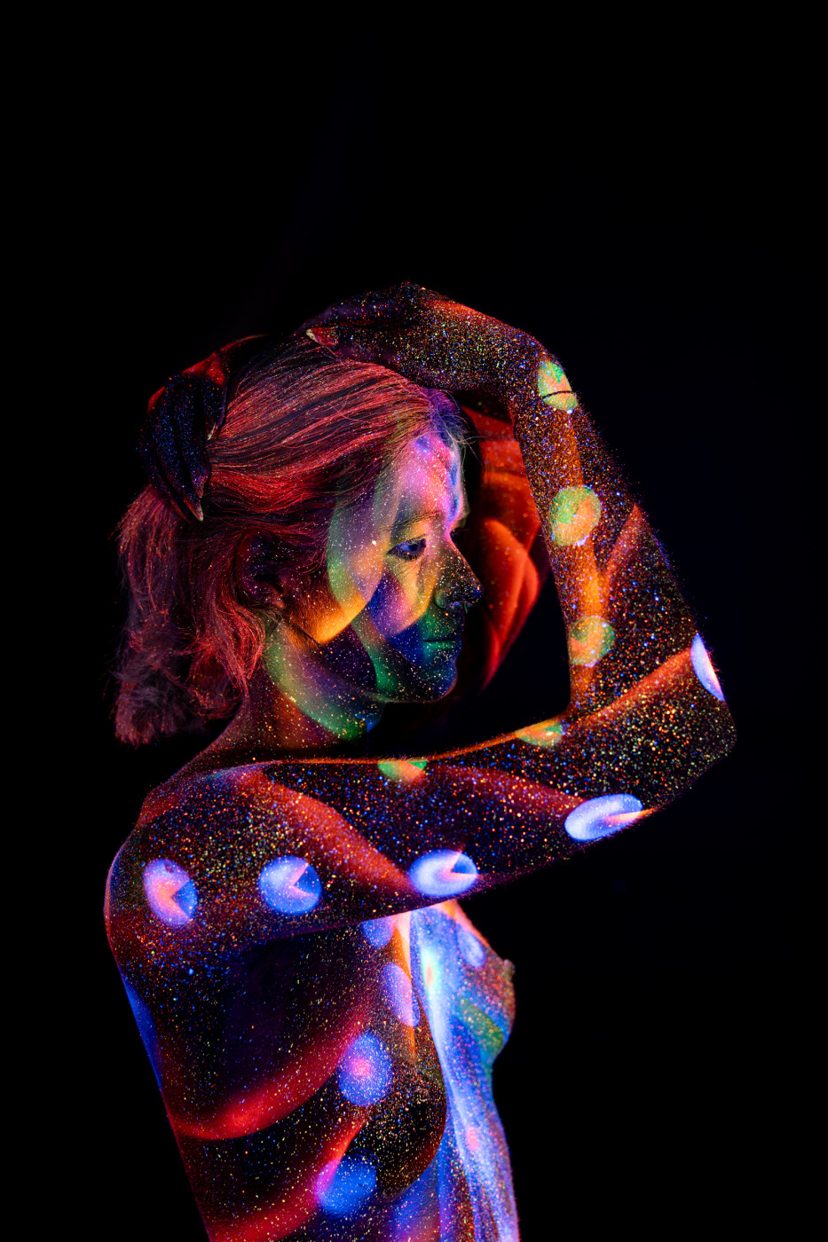 Selina stands in a three-quarter pose, her torso marked with a UV reactive bodypaint pattern of circular dots and linework in red, blue and green. Bodypaint by Elad Itzkin. Photographed by Elad Itzkin Photography under UV blacklight in his studio in Gesundbrunnen, Berlin.