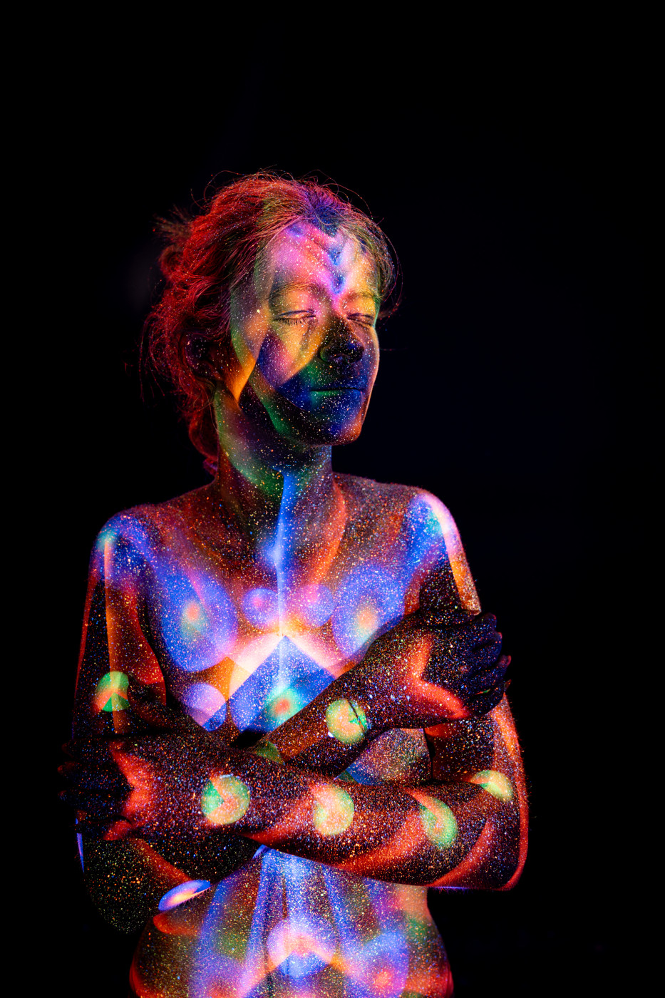 Selina stands in a front facing pose with her head tilted downward. Her torso is painted in a UV reactive design of blue, orange and green polka dots layered over a geometric chevron pattern. Bodypaint by Elad Itzkin. Photographed by Elad Itzkin Photography under UV blacklight in his studio in Gesundbrunnen, Berlin.