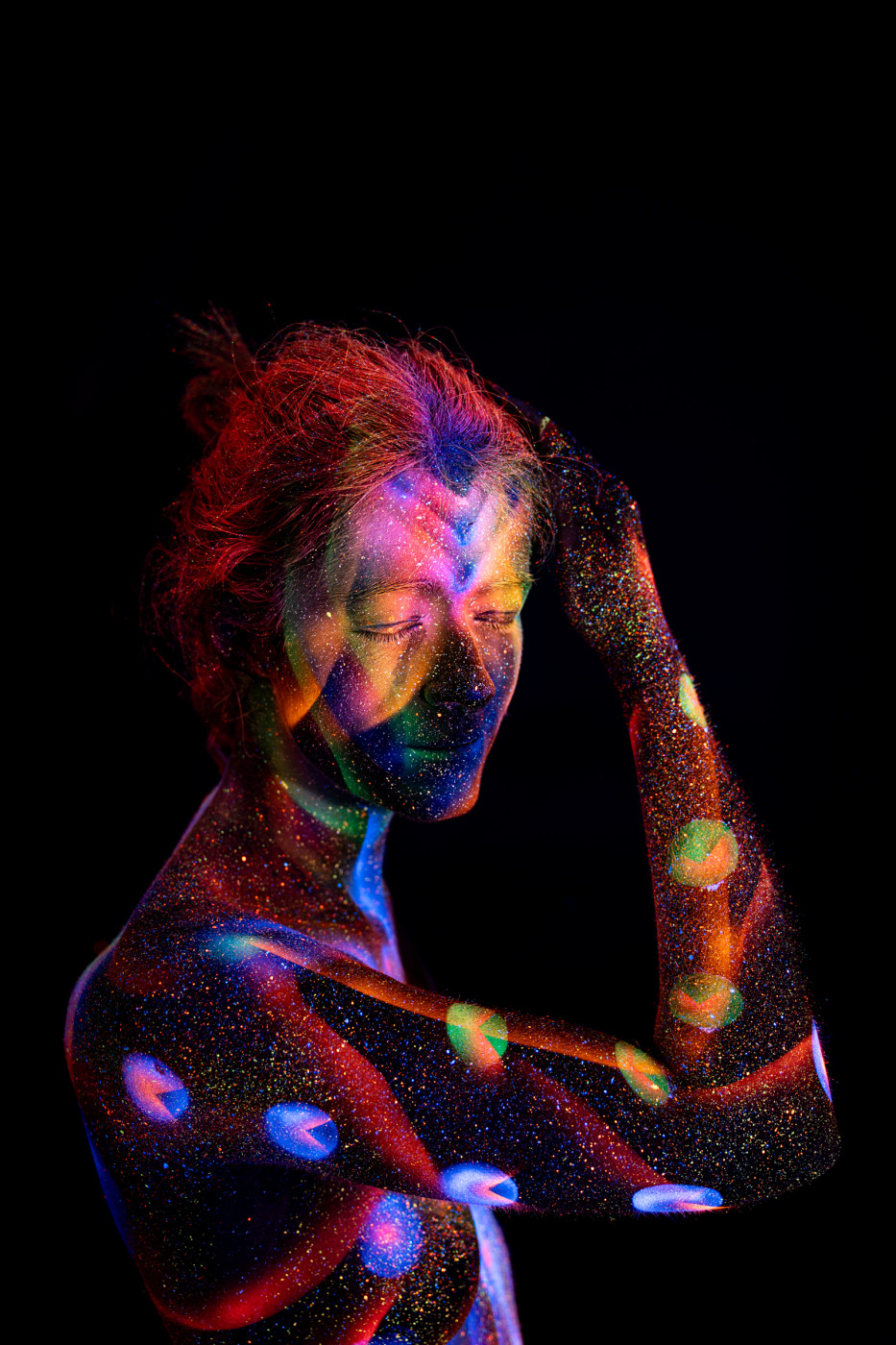 Selina is shown in a side profile, standing with her head bowed slightly forward. The UV reactive bodypaint uses blue, orange and green tones in a pattern of large polka dots across the shoulder and torso. Bodypaint by Elad Itzkin. Photographed by Elad Itzkin Photography under UV blacklight in his studio in Gesundbrunnen, Berlin.