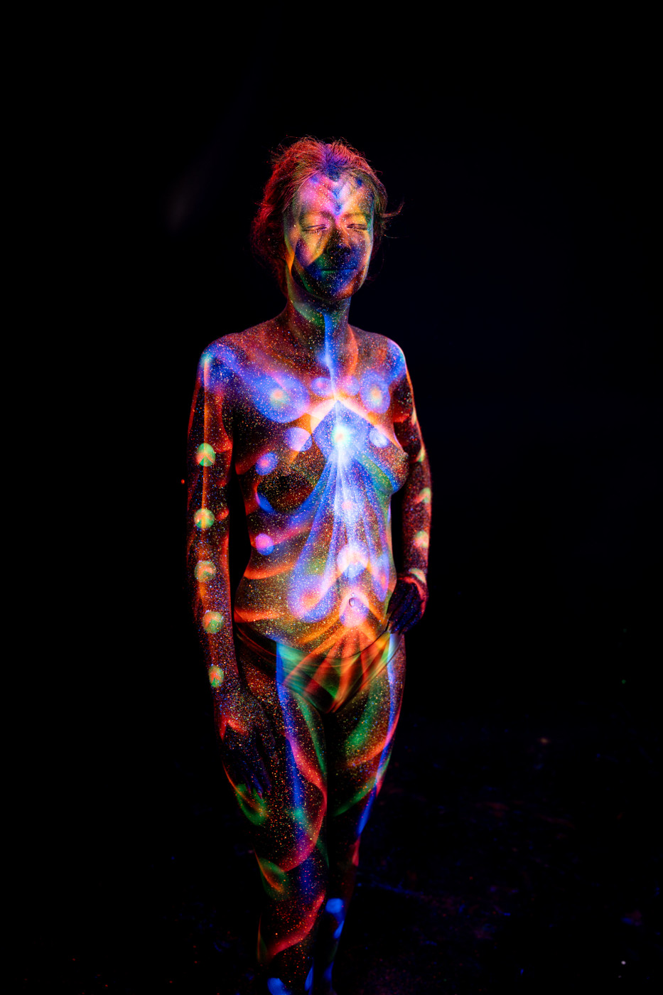 Selina stands facing forward in a full length pose with her head tilted down. The bodypaint covers her torso and legs in a symmetrical geometric design of radiating chevron lines in blue, orange and green, with rows of dots along the sides. Bodypaint by Elad Itzkin. Photographed by Elad Itzkin Photography under UV blacklight in his studio in Gesundbrunnen, Berlin.