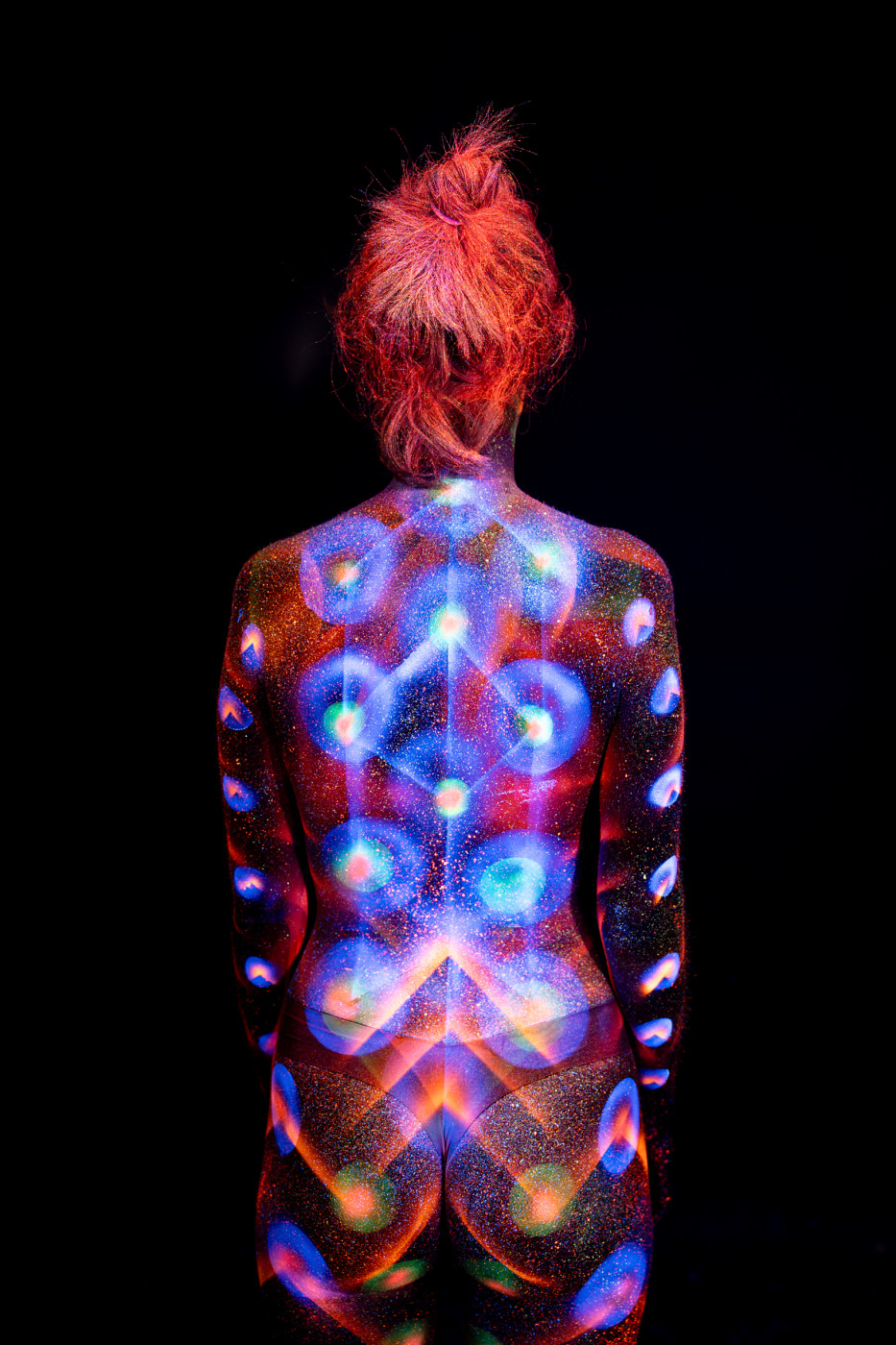 Selina stands with her back to the camera in a full length pose. Her back is painted with a symmetrical UV reactive design of large blue and orange circles arranged in a radial pattern along the spine. Bodypaint by Elad Itzkin. Photographed by Elad Itzkin Photography under UV blacklight in his studio in Gesundbrunnen, Berlin.