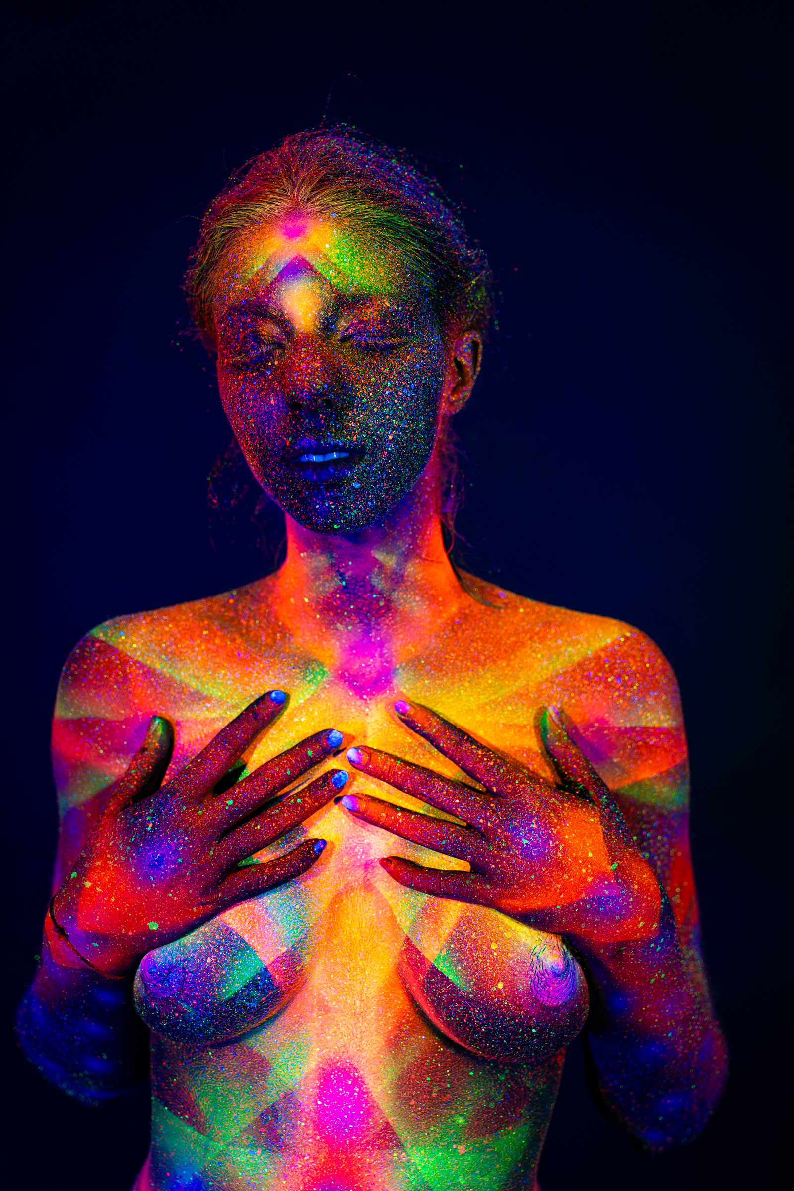 Sofia stands facing forward with her head tilted slightly down. A symmetrical geometric pattern of radiating lines in glowing pink, orange, and green tones covers her chest and shoulders. Bodypaint by Elad Itzkin. Photographed by Elad Itzkin Photography under UV/blacklight in his studio in Gesundbrunnen, Berlin.