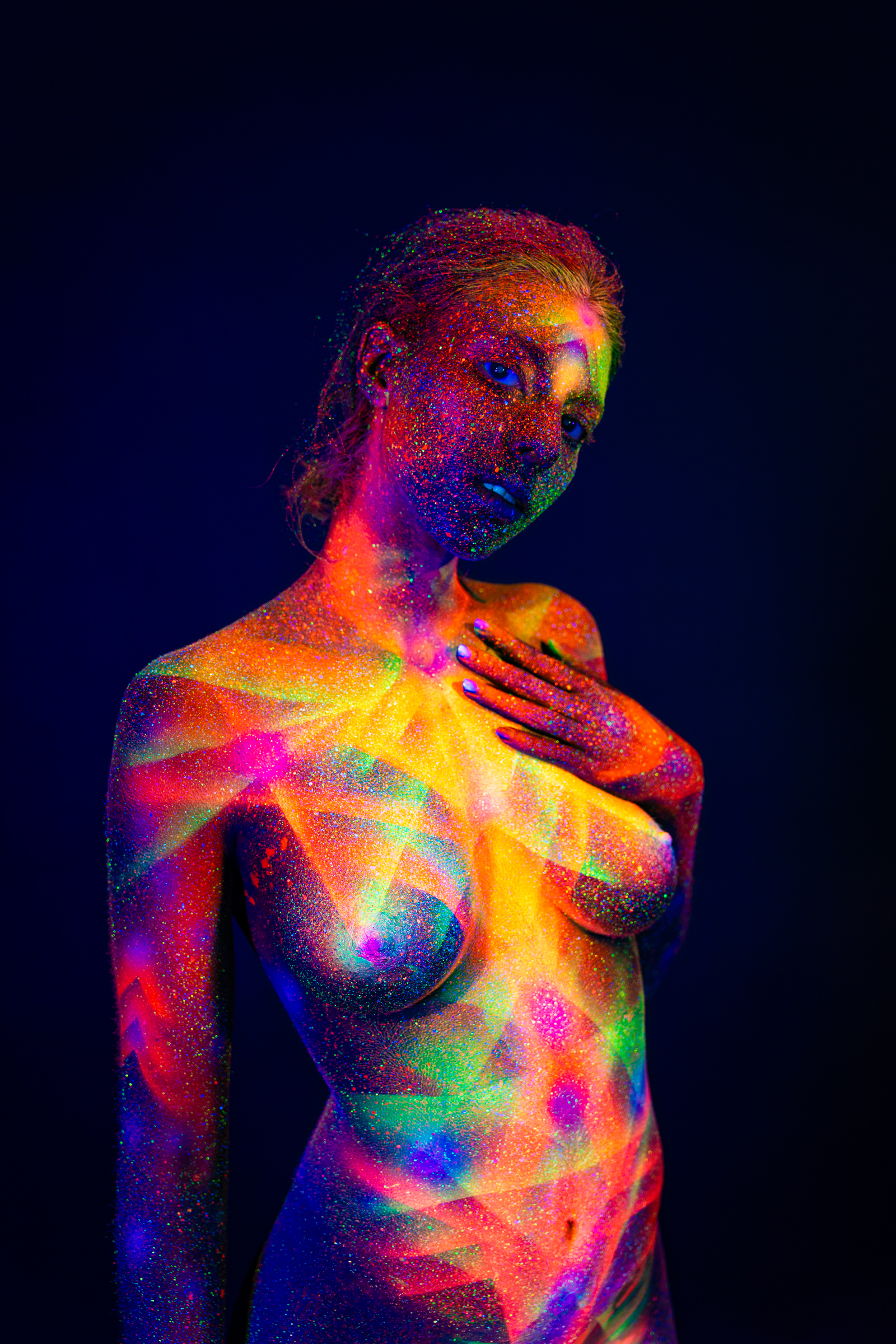 Sofia stands in a three-quarter pose with her head lowered and turned to the side. Her chest and shoulder are decorated with a radiating geometric linework design in UV-reactive colors. Bodypaint by Elad Itzkin. Photographed by Elad Itzkin Photography under UV/blacklight in his studio in Gesundbrunnen, Berlin.