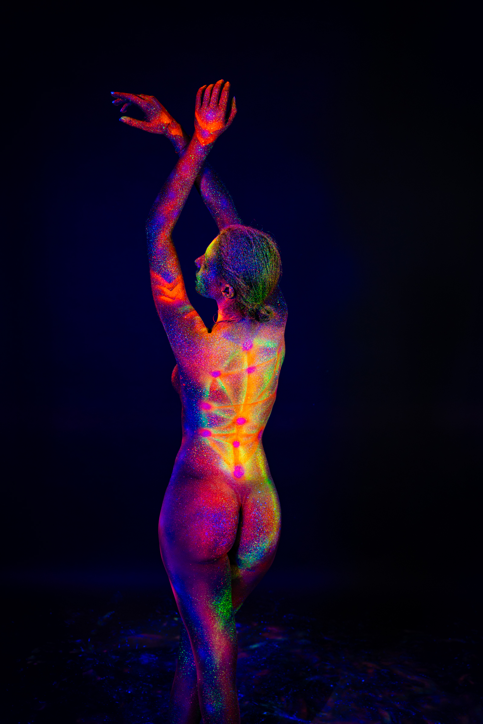 Sofia stands with her back turned to the camera, shown in a full-length pose. A geometric pattern of dots connected by radiating lines glows down the center of her back. Bodypaint by Elad Itzkin. Photographed by Elad Itzkin Photography under UV/blacklight in his studio in Gesundbrunnen, Berlin.