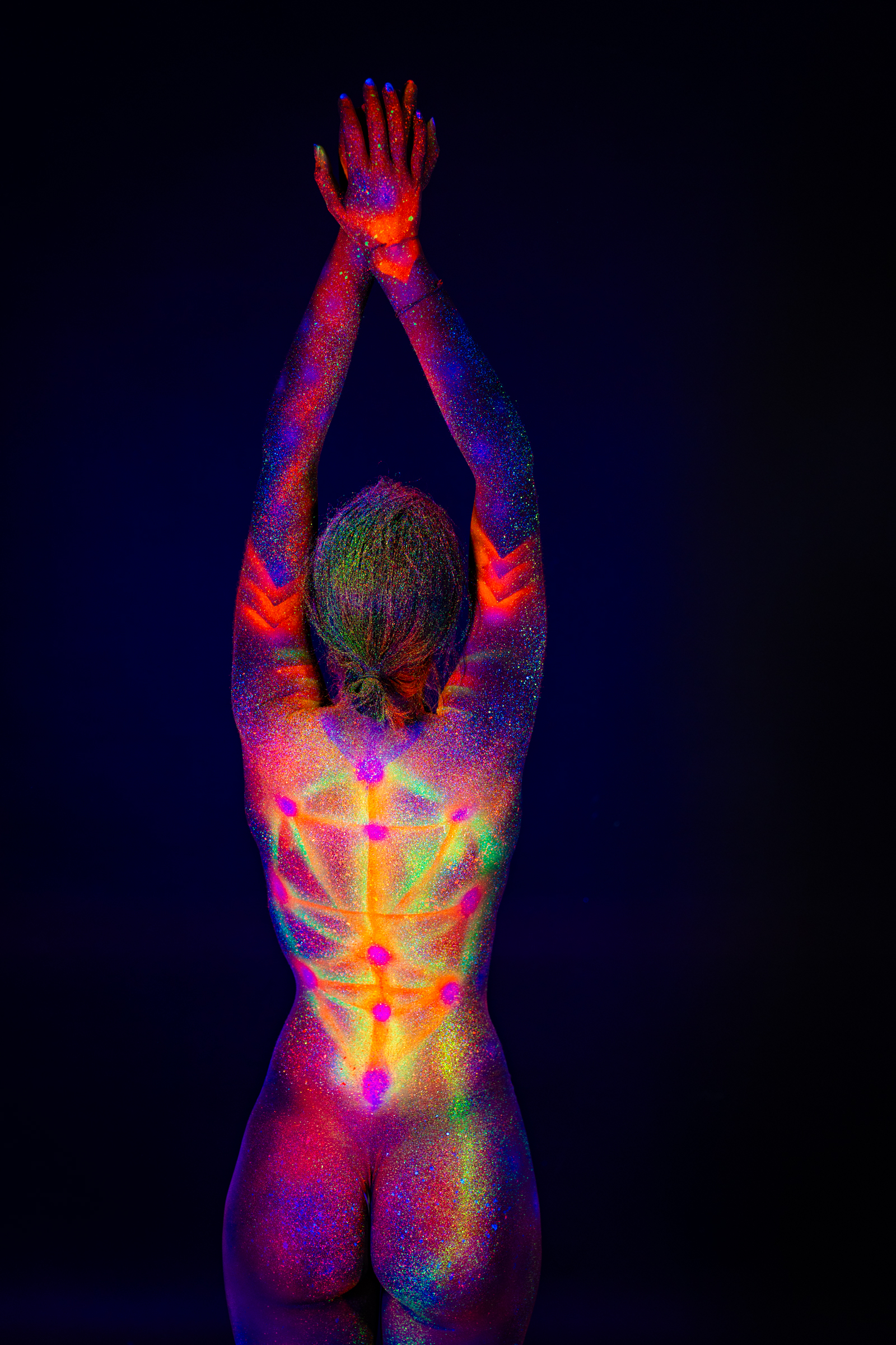 Sofia stands facing away from the camera in a full-length pose. Her spine is marked with a symmetrical geometric design of glowing dots and radiating lines in multiple UV colors. Bodypaint by Elad Itzkin. Photographed by Elad Itzkin Photography under UV/blacklight in his studio in Gesundbrunnen, Berlin.