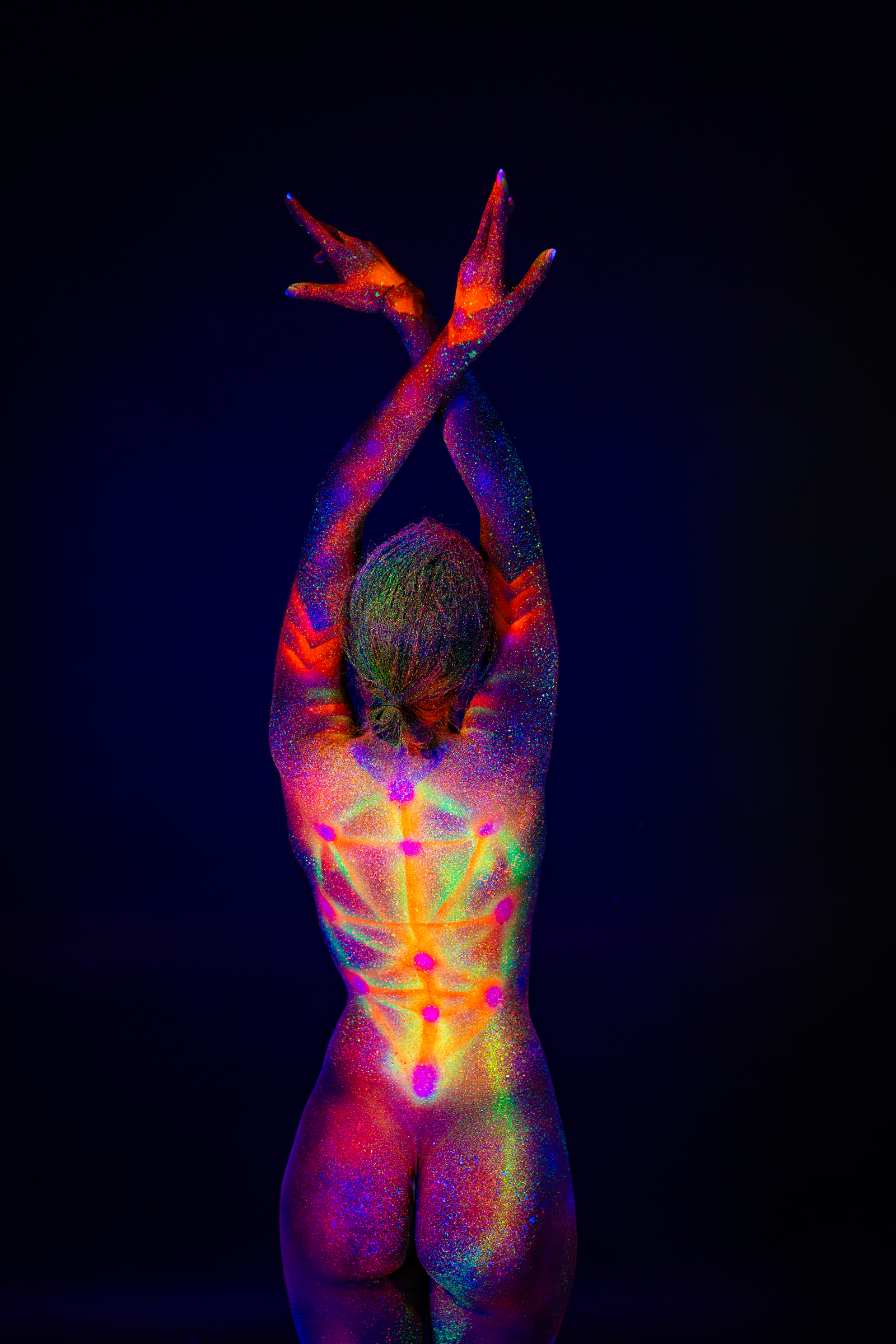 Sofia stands with her back to the camera, shown from head to knees. A rainbow-colored geometric pattern of dots and radiating lines covers her back and spine under blacklight. Bodypaint by Elad Itzkin. Photographed by Elad Itzkin Photography under UV/blacklight in his studio in Gesundbrunnen, Berlin.