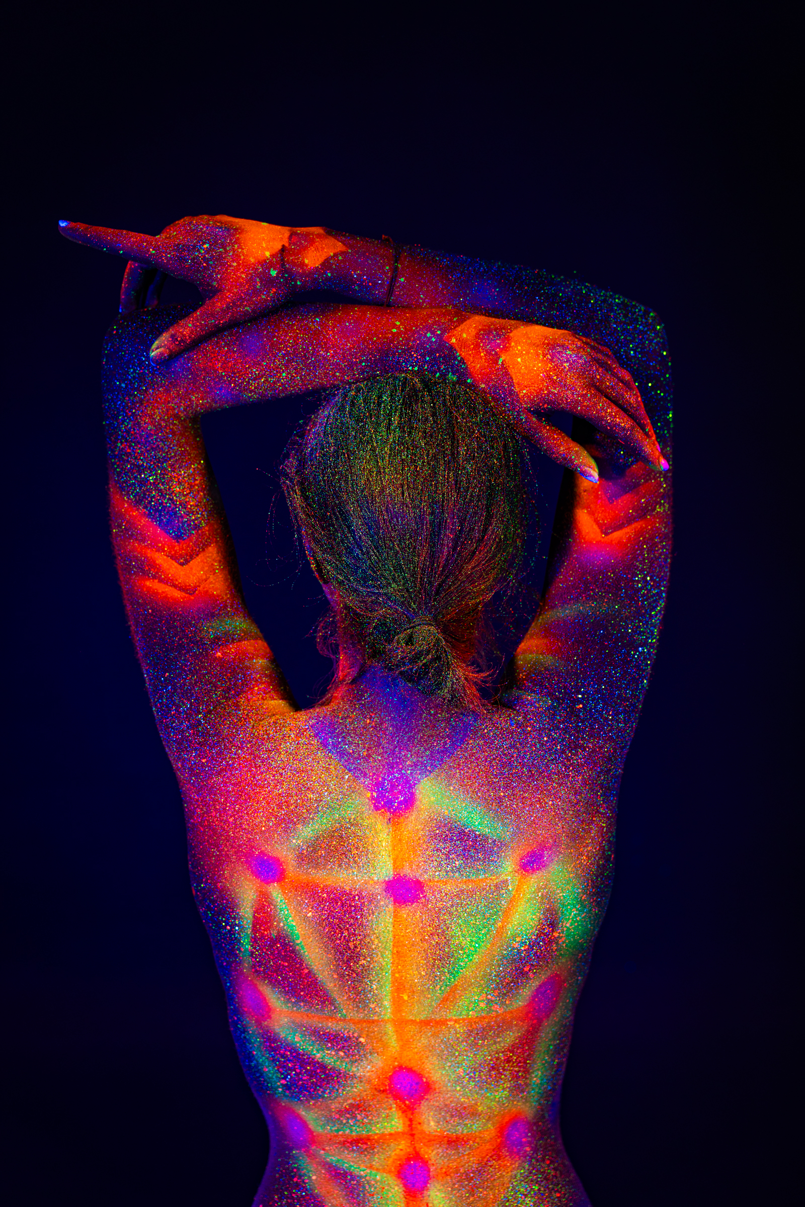 Sofia stands with her back to the camera, framed from the shoulders down. Her upper back is painted with a symmetrical geometric design of dots and radiating lines in UV-reactive color. Bodypaint by Elad Itzkin. Photographed by Elad Itzkin Photography under UV/blacklight in his studio in Gesundbrunnen, Berlin.
