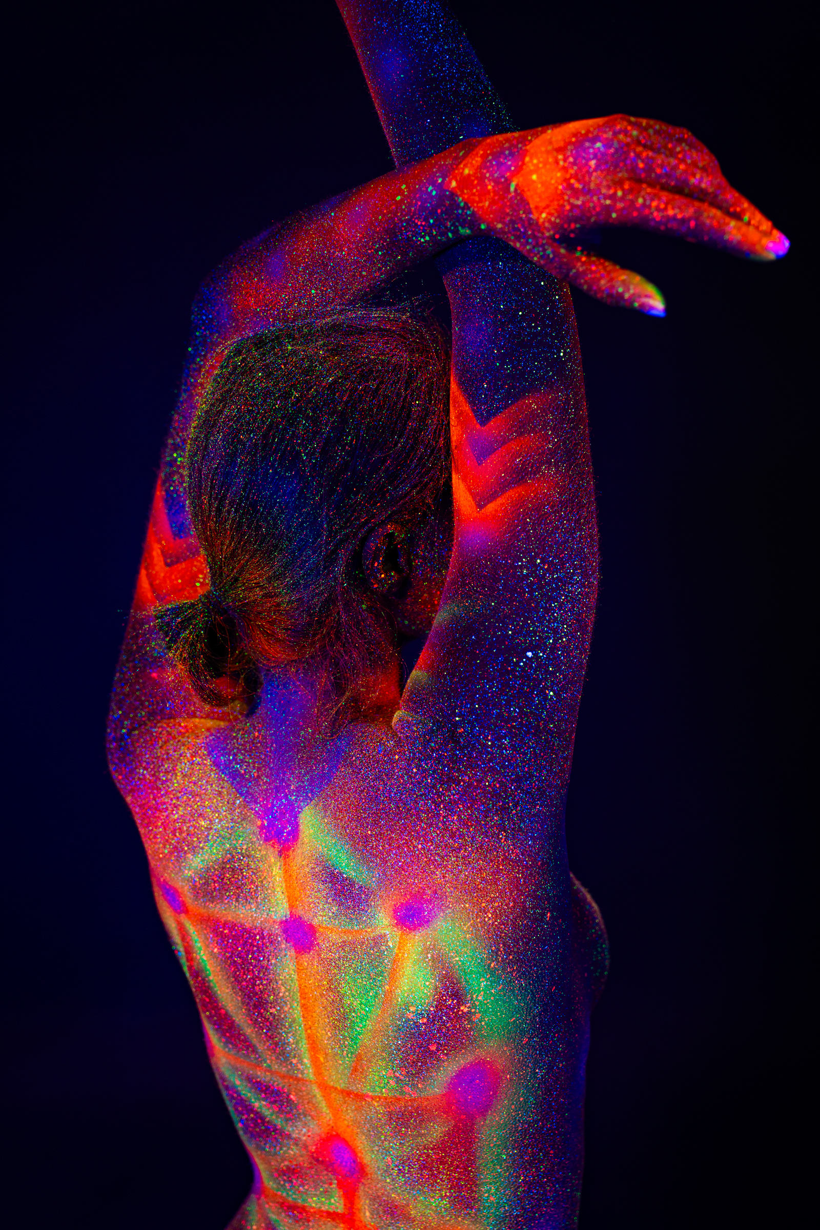 Sofia is photographed from behind in a close crop of her upper back, head tilted forward. A geometric linework design of dots and connecting lines glows in orange, pink, and green under UV light. Bodypaint by Elad Itzkin. Photographed by Elad Itzkin Photography under blacklight in his Berlin studio.