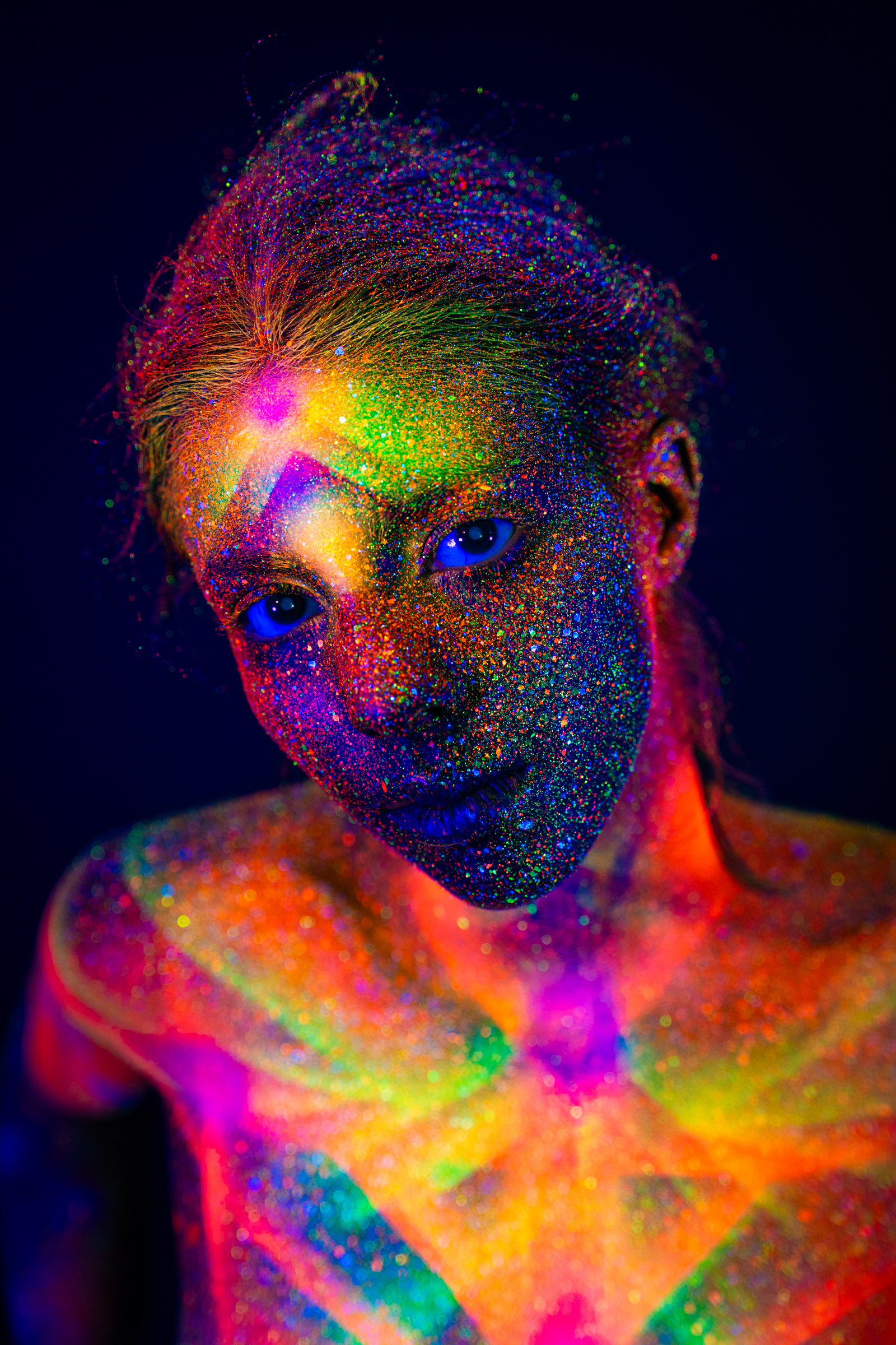 Sofia faces the camera directly in a close portrait. Her face and chest are covered in a radiating geometric pattern in pink, green, yellow, and orange UV pigment. Bodypaint by Elad Itzkin. Photographed by Elad Itzkin Photography under UV light in his Gesundbrunnen, Berlin studio.