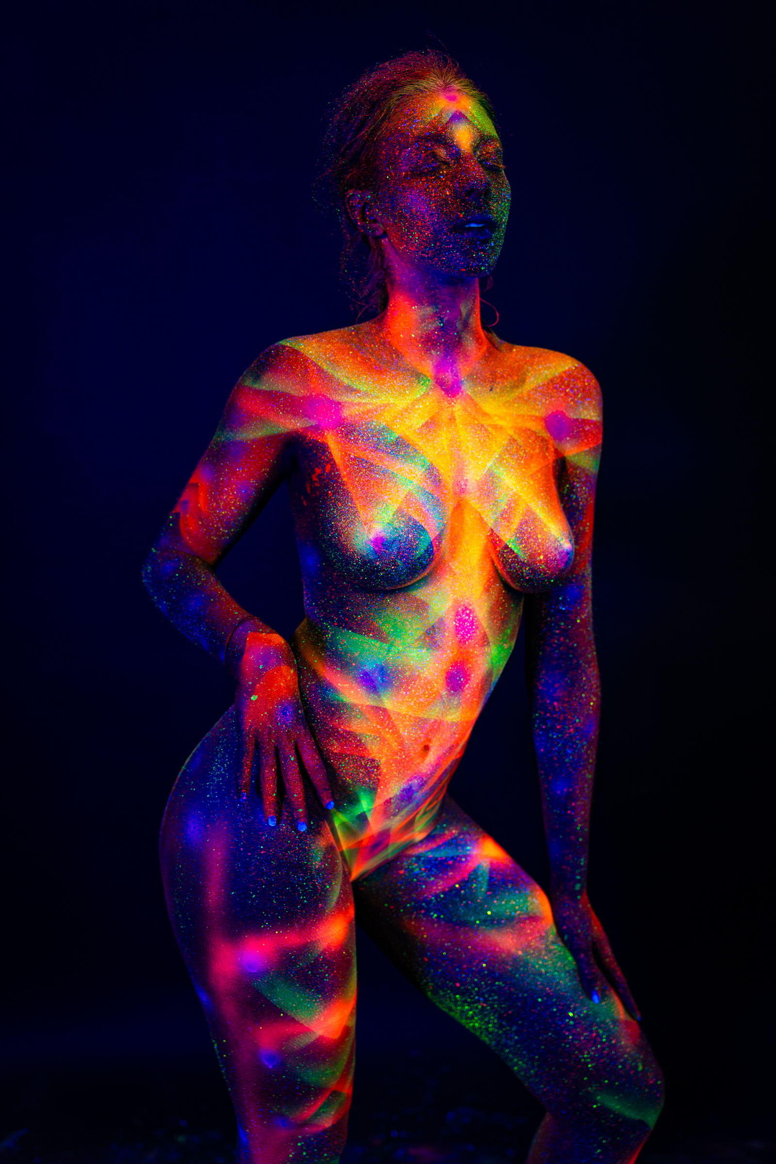 Sofia is photographed in a crouching, three-quarter pose facing the camera. A radiating geometric star pattern in orange, pink, and green covers her chest and torso. Bodypaint by Elad Itzkin. Photographed by Elad Itzkin Photography under UV light in his studio in Gesundbrunnen, Berlin.