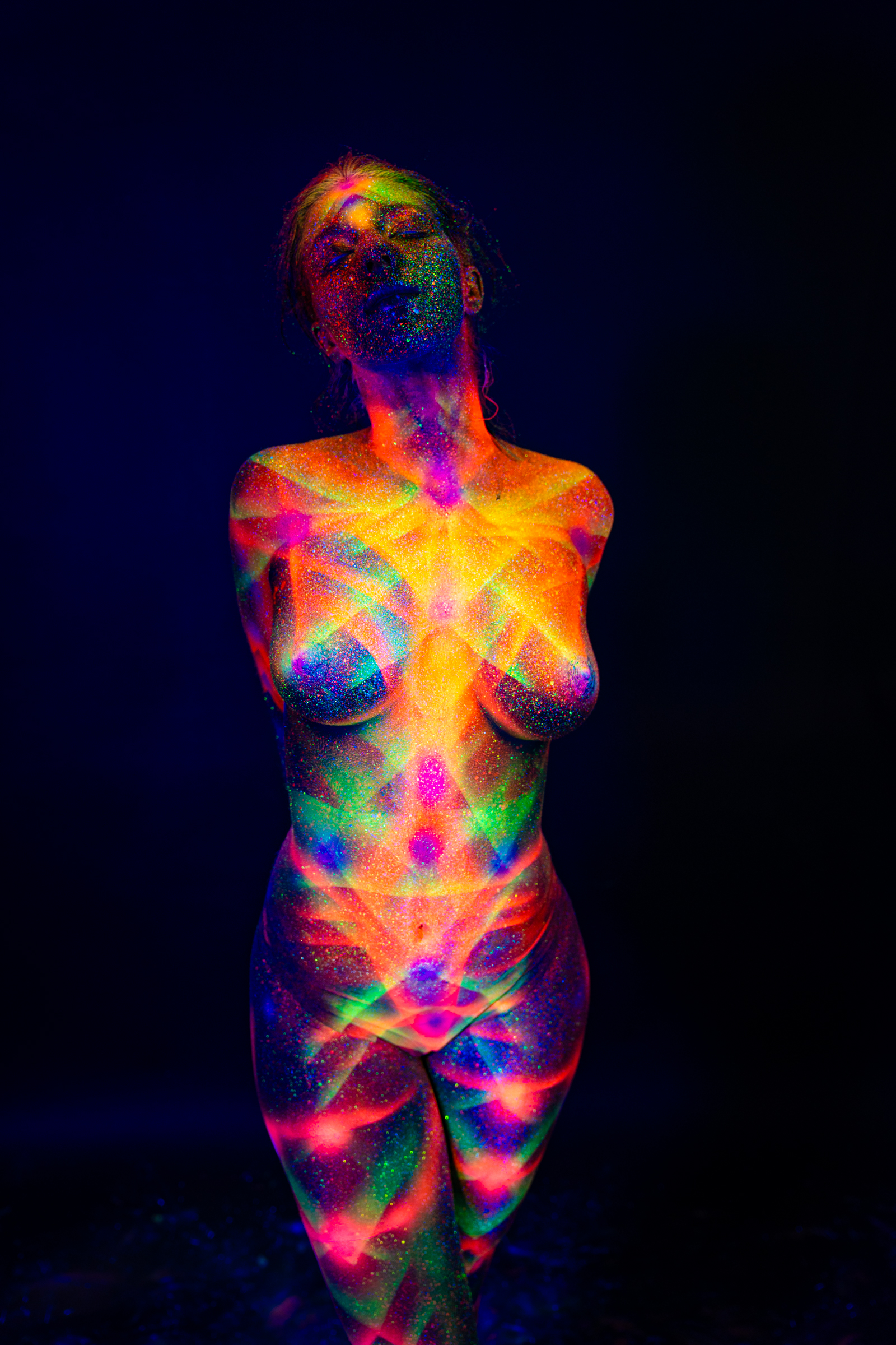 Sofia stands facing forward with her head tilted back. A multicolor geometric pattern in orange, pink, and green UV bodypaint covers her chest, neck, and face. Bodypaint by Elad Itzkin. Photographed by Elad Itzkin Photography under UV/blacklight in his studio in Gesundbrunnen, Berlin.