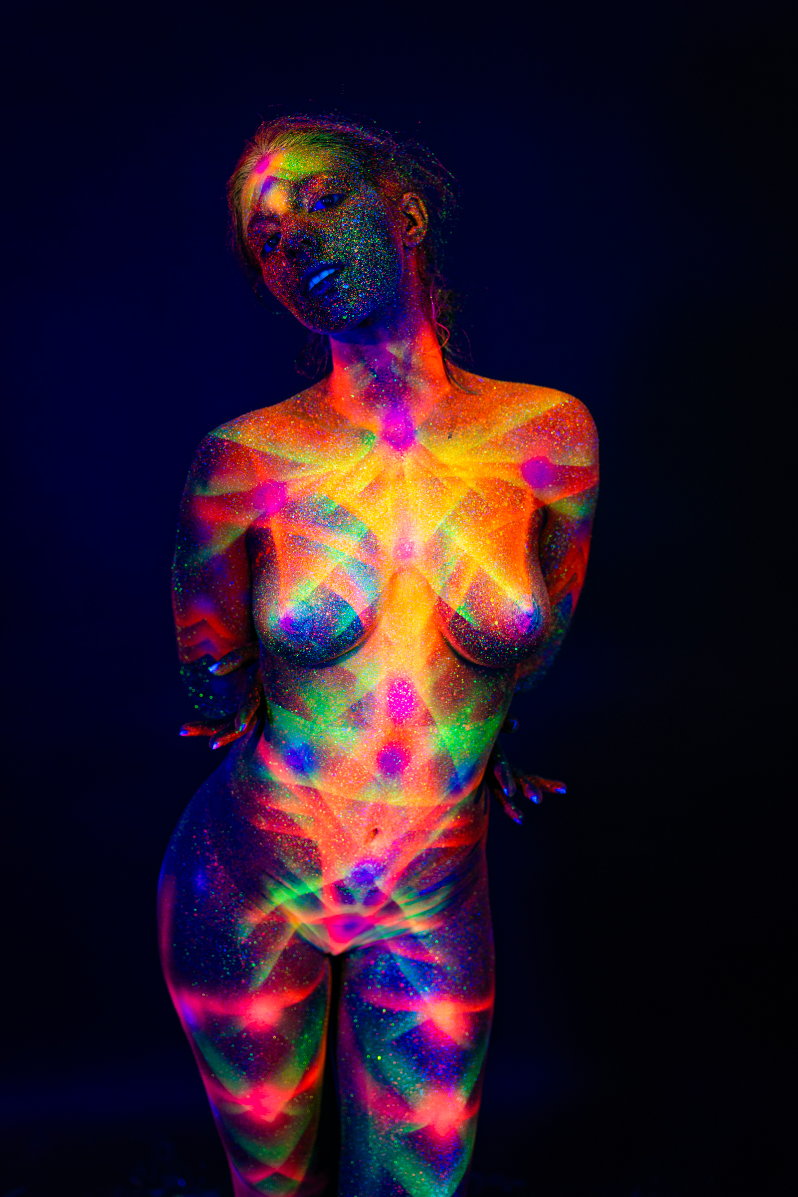 Sofia stands facing forward with her head tilted to one side. A geometric UV bodypaint design in orange, pink, green, and blue covers her torso and face. Bodypaint by Elad Itzkin. Photographed by Elad Itzkin Photography under UV/blacklight in his studio in Gesundbrunnen, Berlin.