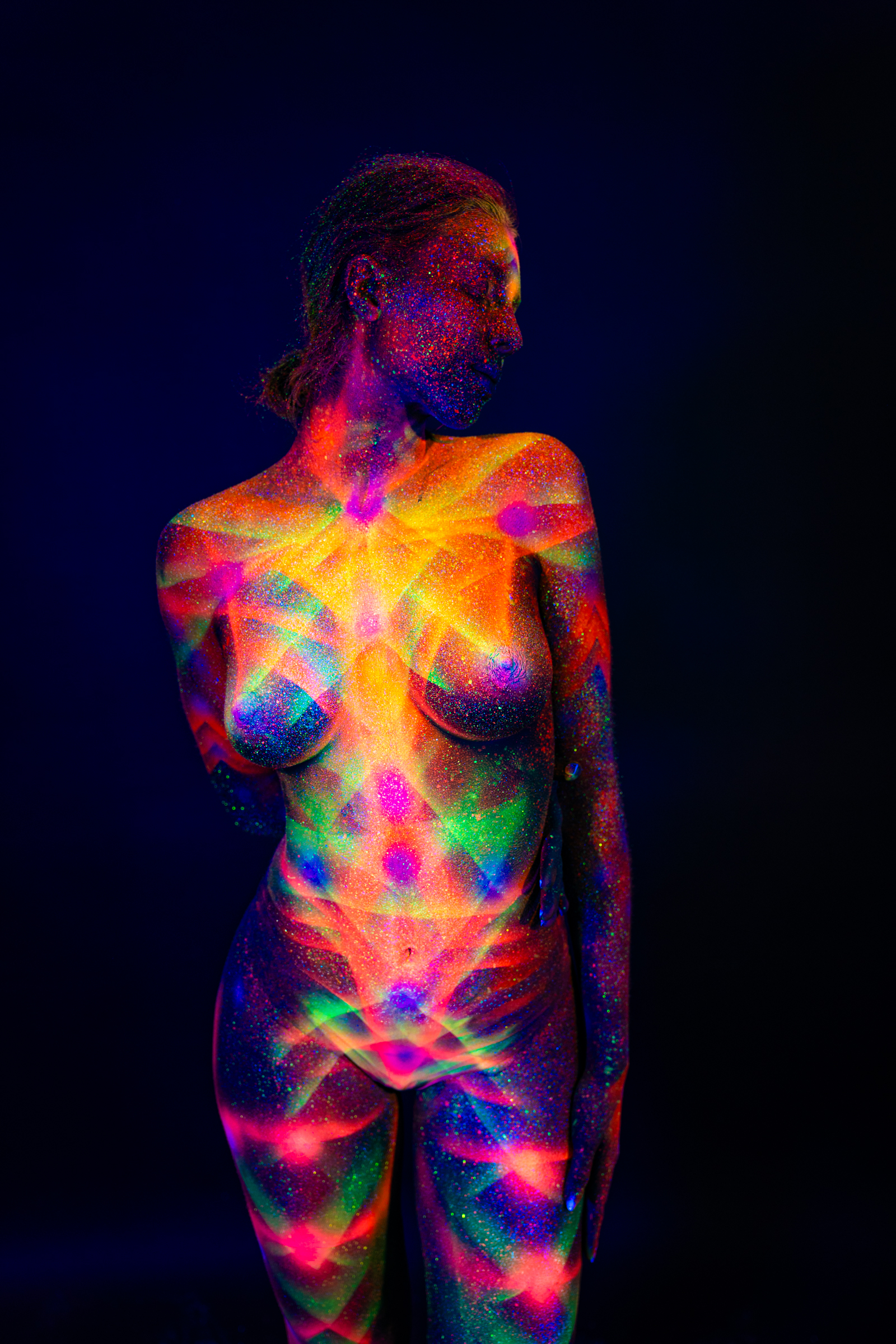 Sofia stands with her torso turned slightly and her head tilted downward. A geometric bodypaint design combining chevron patterns and radiating lines in orange, pink, green, and blue UV pigment covers her chest and torso. Bodypaint by Elad Itzkin. Photographed by Elad Itzkin Photography under UV/blacklight in his studio in Gesundbrunnen, Berlin.