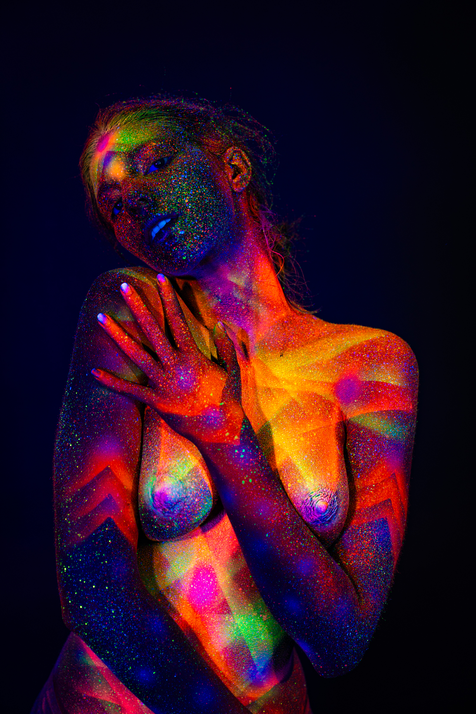 Standing and facing forward, Sofia is painted with a radiating geometric UV bodypaint design in orange, pink, and green across her chest, neck, and face. Bodypaint by Elad Itzkin. Photographed by Elad Itzkin Photography under UV/blacklight in his studio in Gesundbrunnen, Berlin.