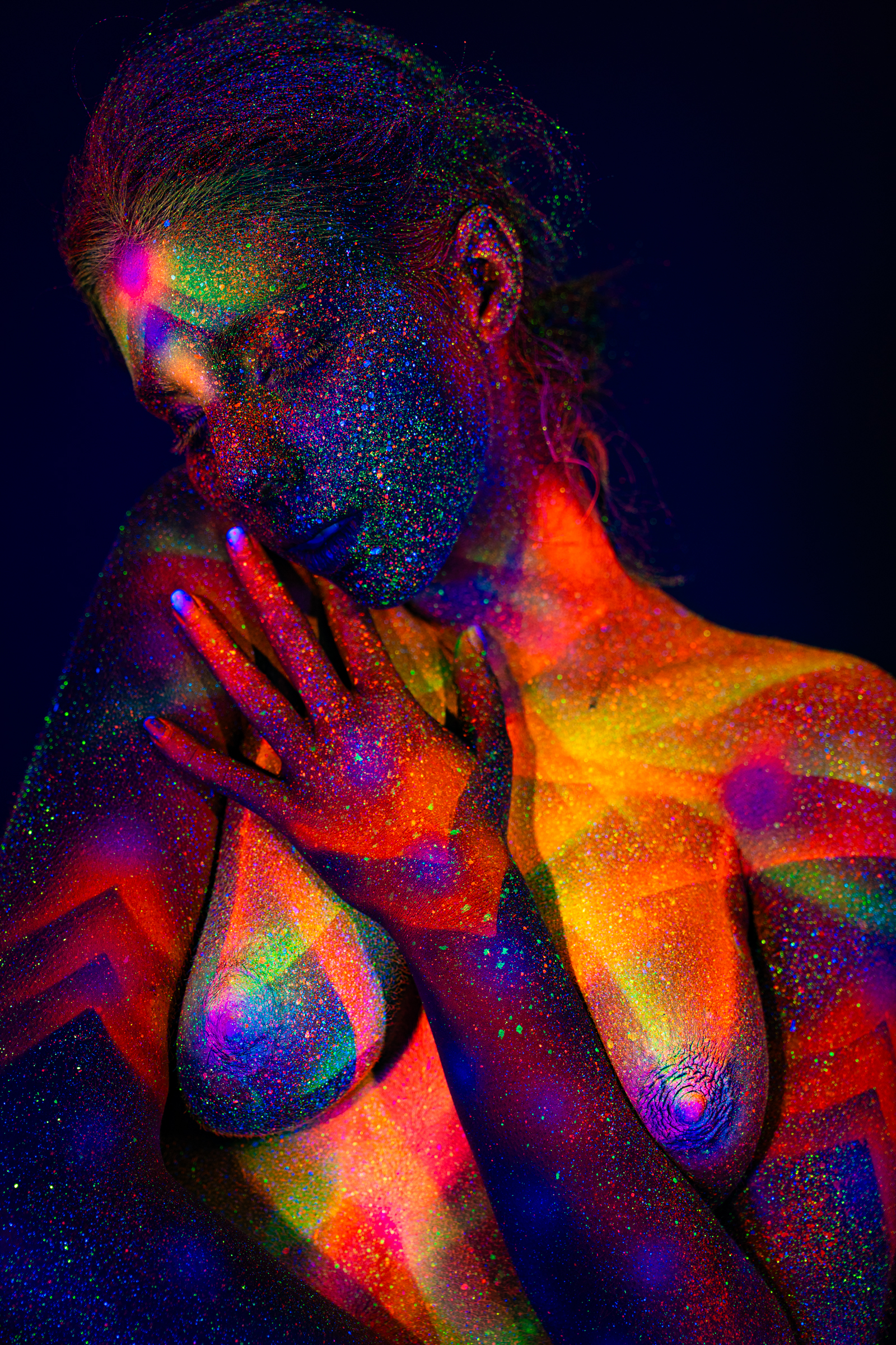 Sofia is shown in an upper-body portrait, standing with her head tilted to one side. Orange, pink, and green UV bodypaint forms a chevron and geometric radiating design across her torso and shoulder. Bodypaint by Elad Itzkin. Photographed by Elad Itzkin Photography under UV/blacklight in his studio in Gesundbrunnen, Berlin.