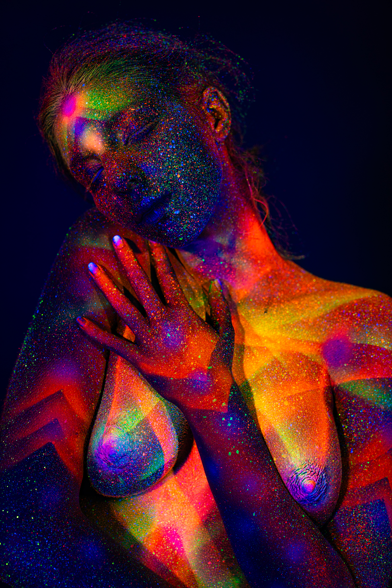In an upper-body portrait, Sofia stands with her head tilted to one side. A chevron and radiating geometric bodypaint pattern in orange, pink, and green UV pigment covers her torso and shoulder. Bodypaint by Elad Itzkin. Photographed by Elad Itzkin Photography under UV/blacklight in his studio in Gesundbrunnen, Berlin.