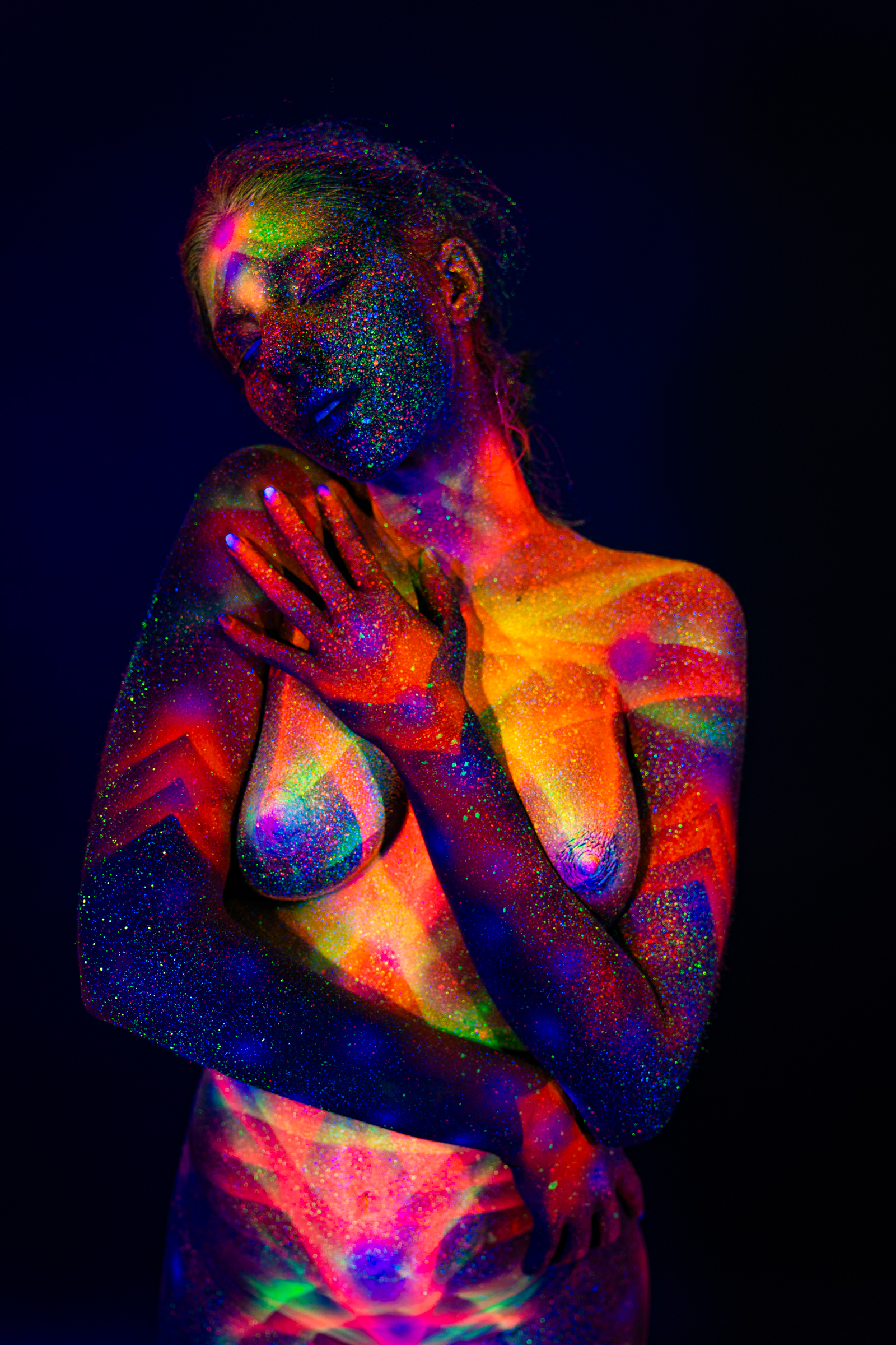 Sofia stands in an upper-body portrait, her torso turned slightly to one side. A geometric bodypaint design combining chevrons and radiating lines in orange, pink, and green UV pigment covers her chest. Bodypaint by Elad Itzkin. Photographed by Elad Itzkin Photography under UV/blacklight in his studio in Gesundbrunnen, Berlin.