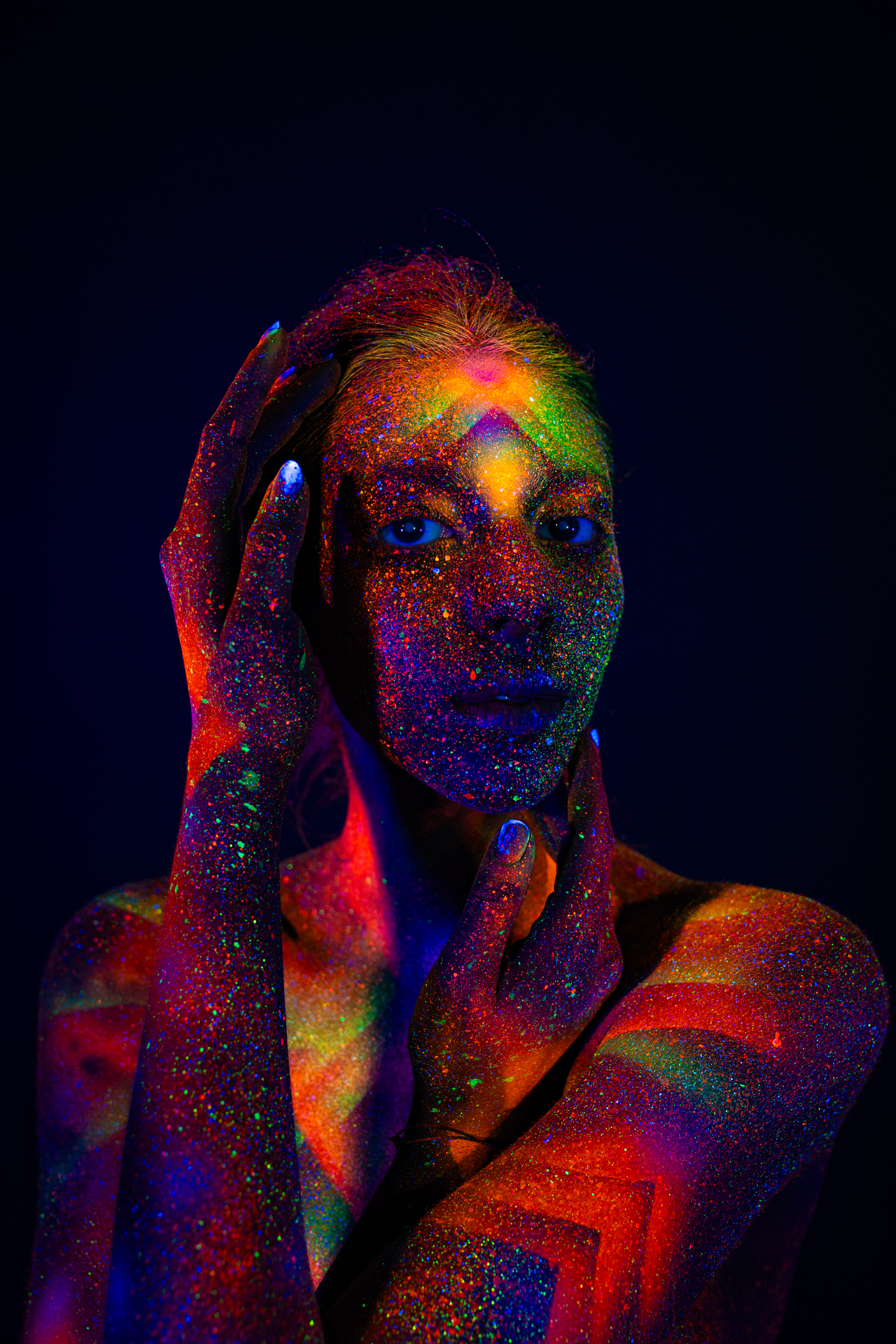 Standing and facing the camera in a close upper-body portrait, Sofia wears a radiating geometric bodypaint pattern in orange, pink, and green UV pigment across her face and chest. Bodypaint by Elad Itzkin. Photographed by Elad Itzkin Photography under UV/blacklight in his studio in Gesundbrunnen, Berlin.