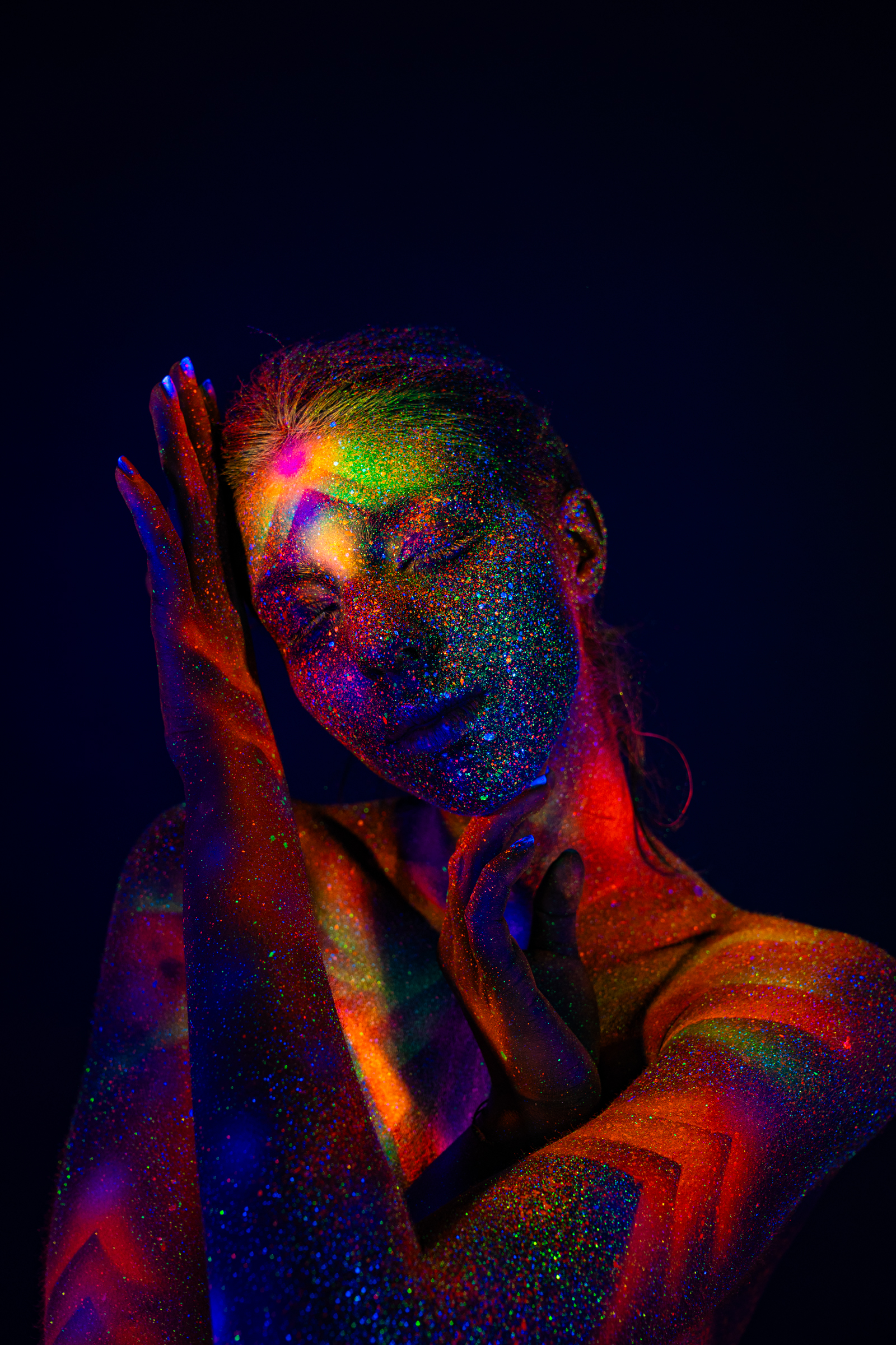In a close upper-body portrait, Sofia stands with her head tilted to one side. A radiating geometric bodypaint pattern in orange, pink, and green UV pigment covers her face, neck, and shoulders. Bodypaint by Elad Itzkin. Photographed by Elad Itzkin Photography under UV/blacklight in his studio in Gesundbrunnen, Berlin.