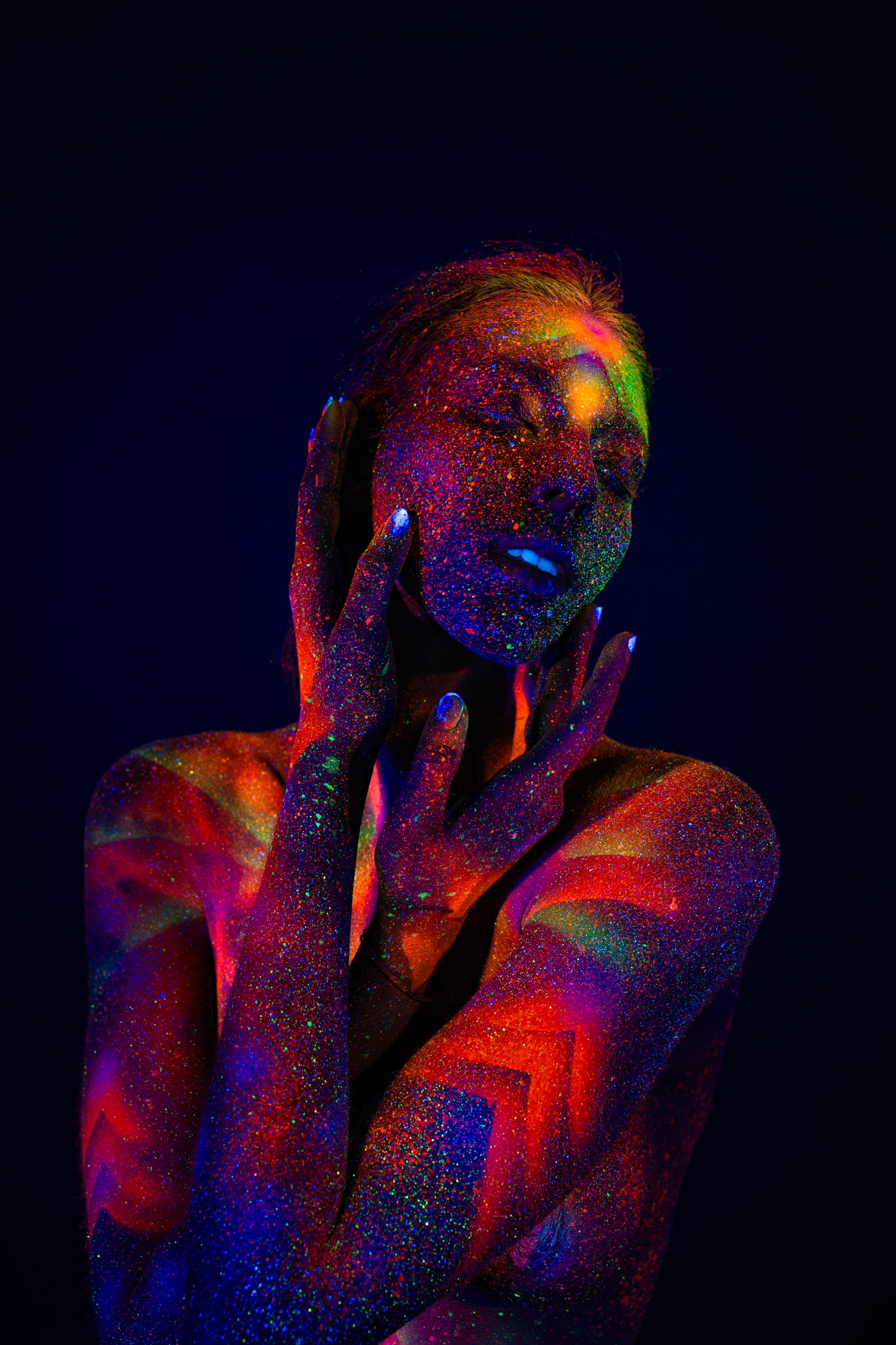 Sofia is shown in an upper-body portrait with her head tilted back and to the side. A geometric radiating bodypaint design in orange, pink, and green UV pigment covers her face and chest. Bodypaint by Elad Itzkin. Photographed by Elad Itzkin Photography under UV/blacklight in his studio in Gesundbrunnen, Berlin.