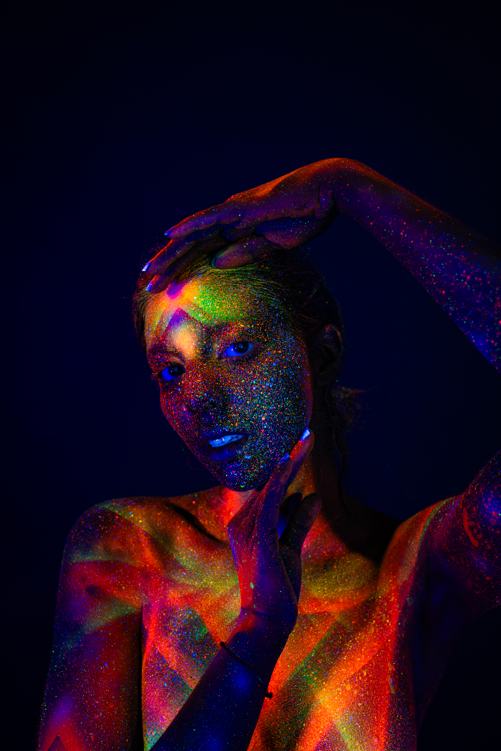 Sofia is photographed facing the camera in an upper-body portrait. A radiating geometric bodypaint design in orange, pink, and green UV pigment covers her face, neck, and torso. Bodypaint by Elad Itzkin. Photographed by Elad Itzkin Photography under UV/blacklight in his studio in Gesundbrunnen, Berlin.