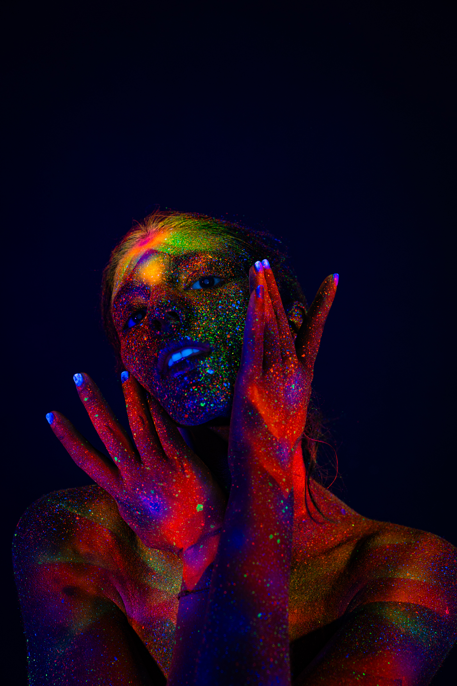 In a close upper-body portrait facing the camera, Sofia wears a radiating geometric UV bodypaint pattern in orange, pink, and green covering her face and shoulders. Bodypaint by Elad Itzkin. Photographed by Elad Itzkin Photography under UV/blacklight in his studio in Gesundbrunnen, Berlin.