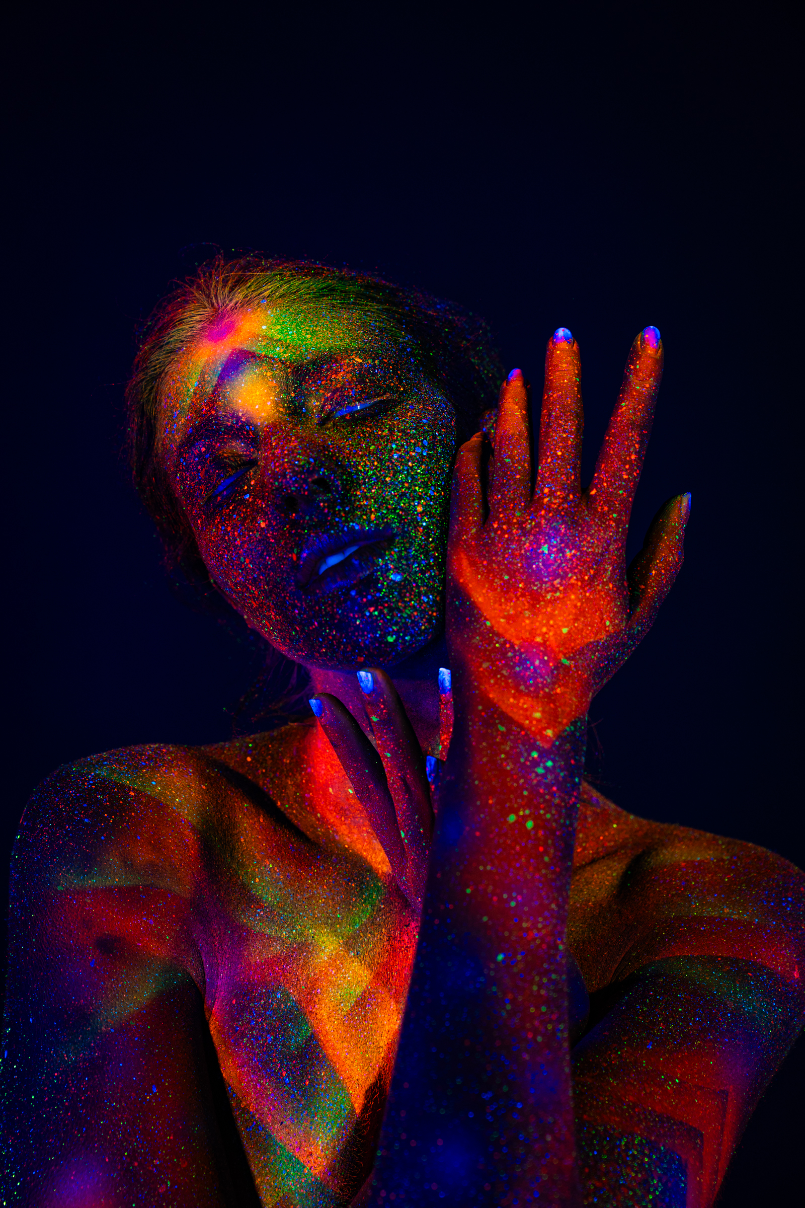 Sofia is shown from the shoulders up, head tilted to one side with her eyes closed. Her skin is covered in a UV-reactive chevron and star pattern in green, orange, and blue tones. Bodypaint by Elad Itzkin. Photographed by Elad Itzkin Photography under blacklight in his studio in Gesundbrunnen, Berlin.