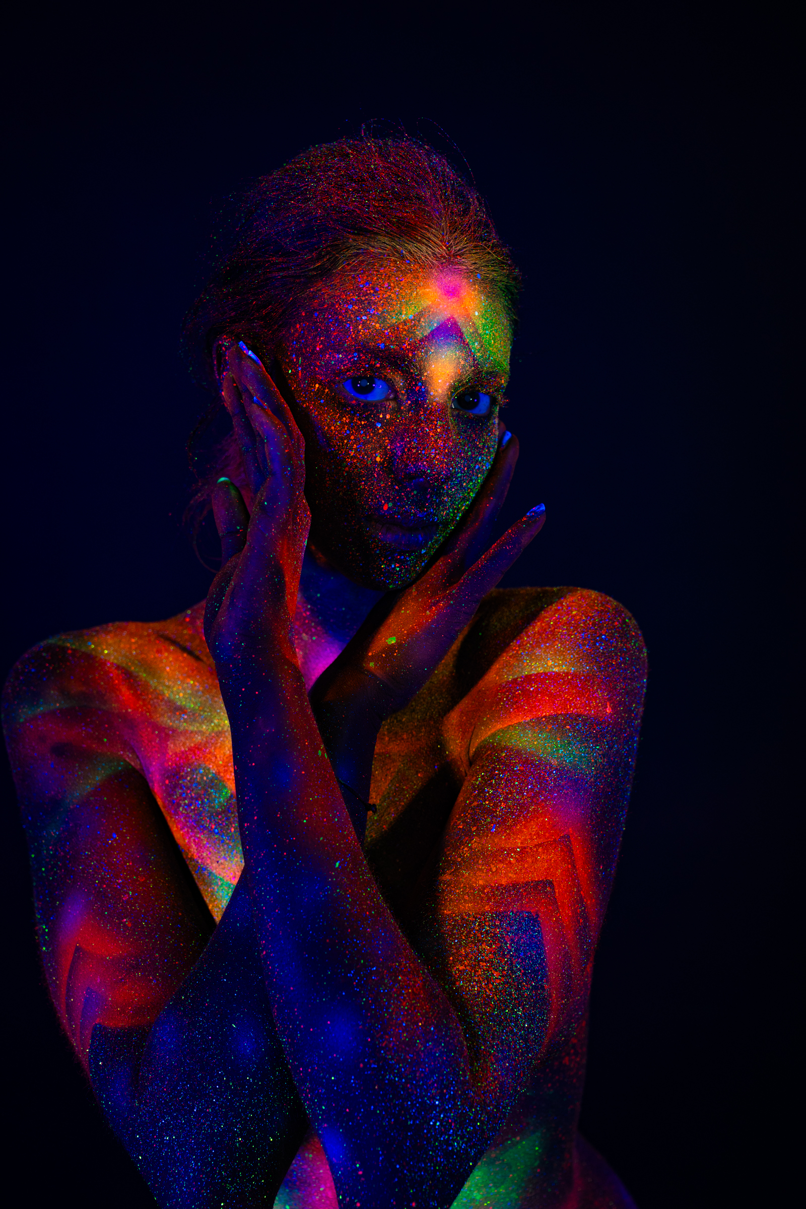 Sofia is photographed in an upper-body portrait, facing the camera with her eyes open. Her face and shoulders are covered in a UV-reactive geometric design in orange, green, and blue glitter tones. Bodypaint by Elad Itzkin. Photographed by Elad Itzkin Photography under UV light in his studio in Gesundbrunnen, Berlin.