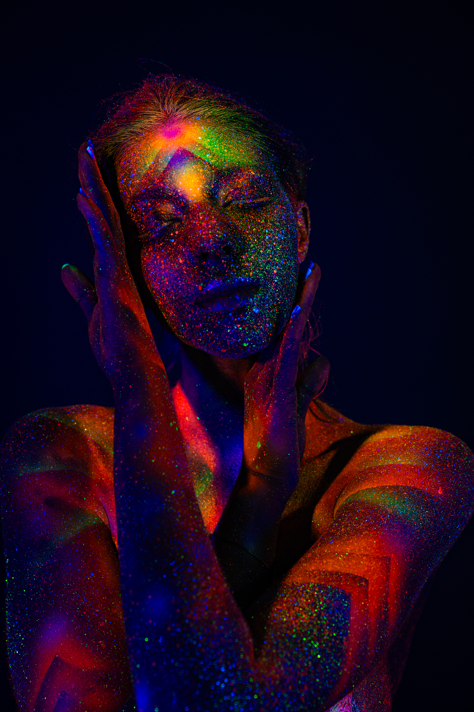 In this upper-body portrait, Sofia's head is tilted back slightly with her eyes closed. A radiating geometric pattern in orange, green, and blue UV paint covers her face, neck, and shoulders. Bodypaint by Elad Itzkin. Photographed under blacklight by Elad Itzkin Photography in his studio in Gesundbrunnen, Berlin.