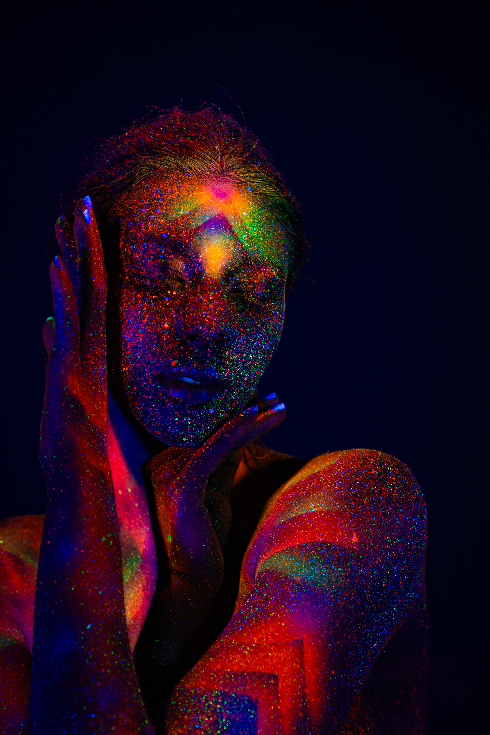 Sofia appears in a close upper-body portrait with her head turned to the side, eyes closed. Her face and shoulders carry a UV-reactive geometric pattern in orange, green, and blue tones. Bodypaint by Elad Itzkin. Photographed by Elad Itzkin Photography under UV light in his studio in Gesundbrunnen, Berlin.
