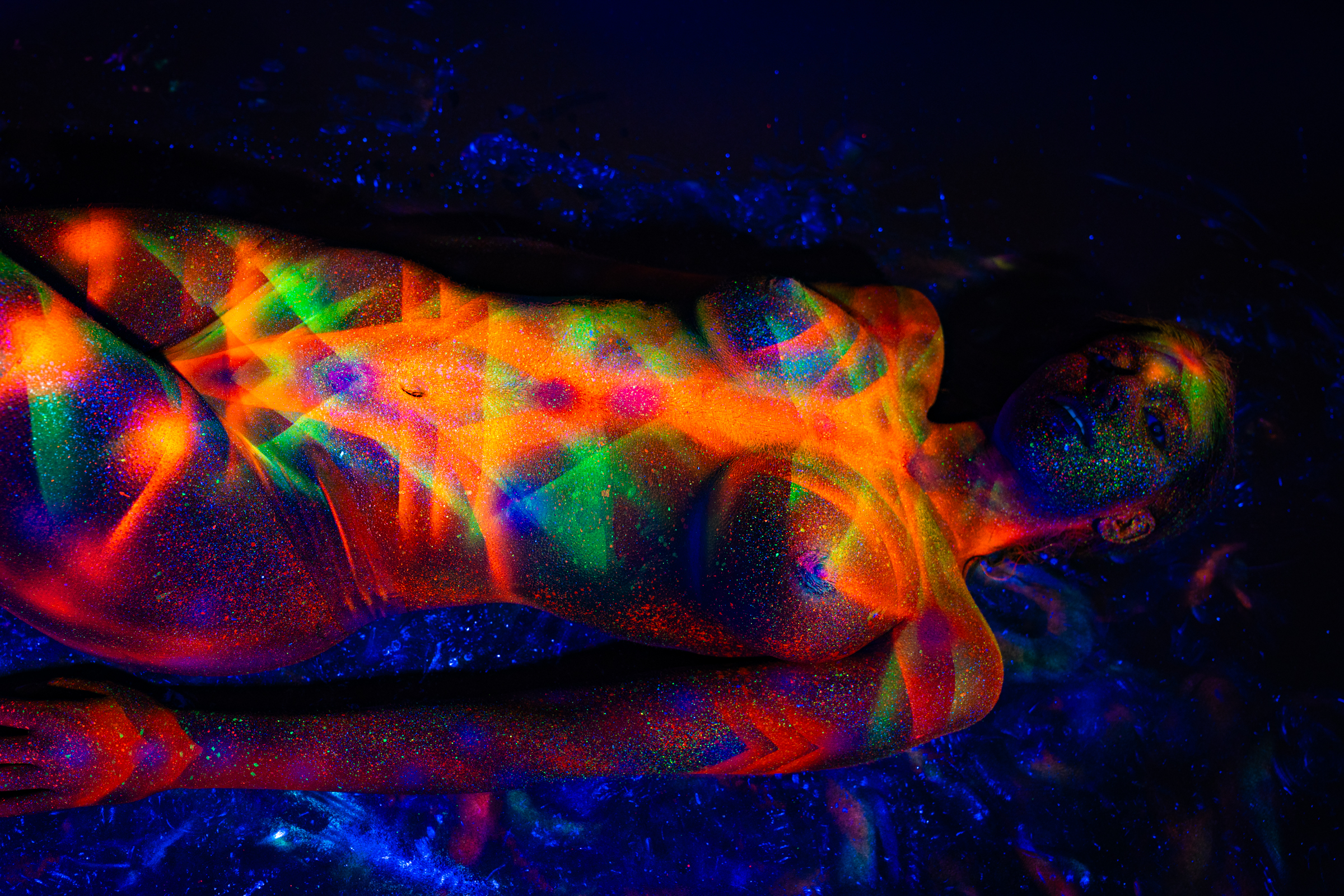 Sofia lies on her back on a reflective surface, viewed from above with her head turned to one side. Her body is covered in a UV-reactive geometric triangle pattern in orange, red, and green. Bodypaint by Elad Itzkin. Photographed by Elad Itzkin Photography under UV light in his Gesundbrunnen, Berlin studio.