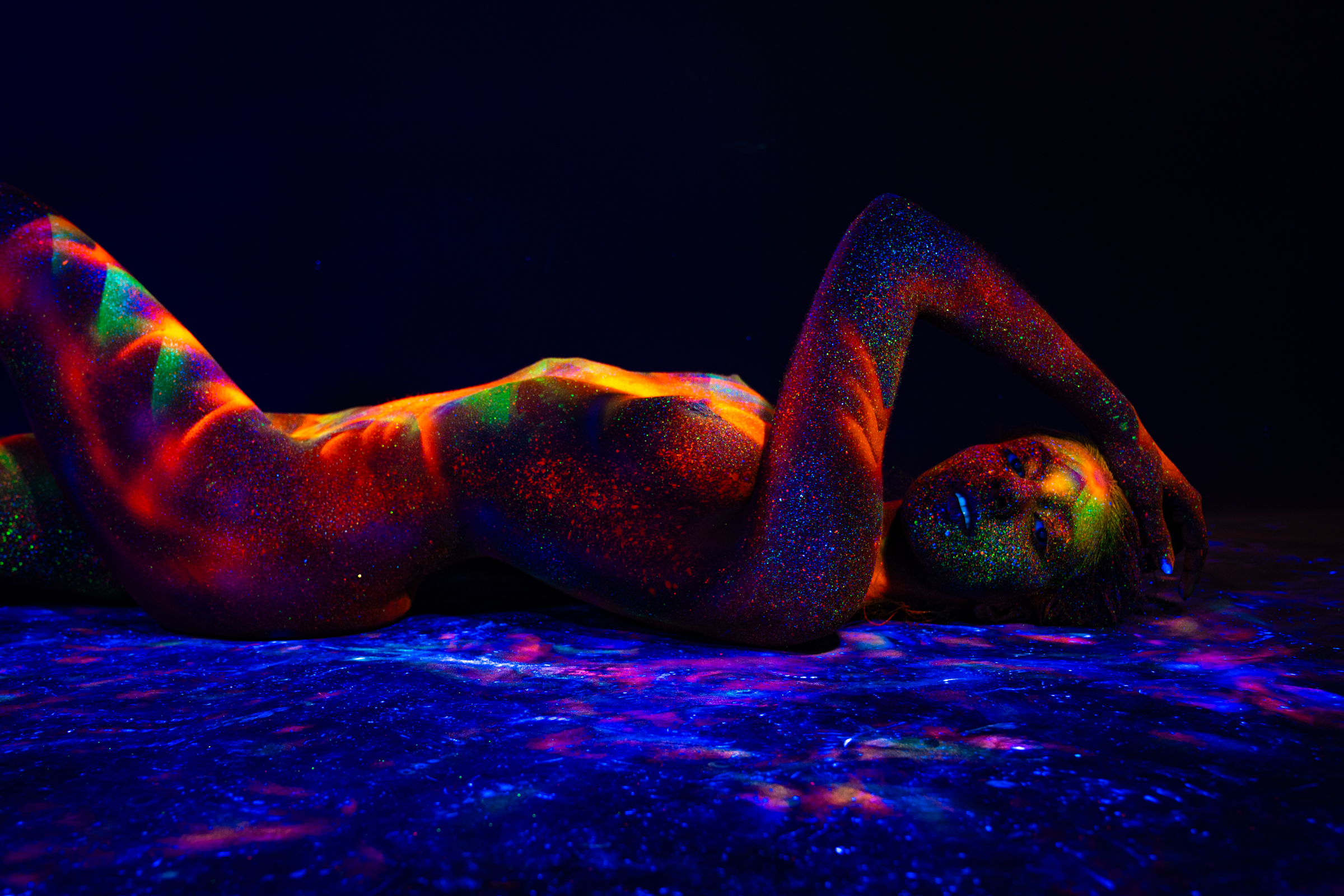 Sofia reclines on her back with her upper body arched and her head tilted back on a reflective surface. A UV-reactive geometric pattern in orange, red, and green covers her torso. Bodypaint by Elad Itzkin. Photographed under blacklight by Elad Itzkin Photography in his studio in Gesundbrunnen, Berlin.