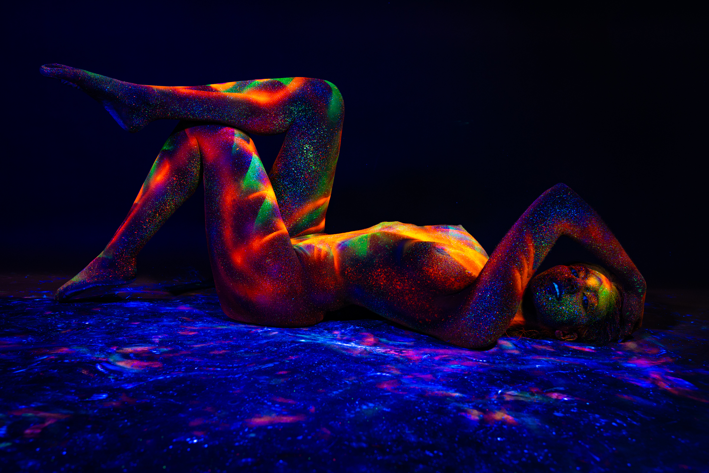 Sofia lies on her back with her knees bent and one leg raised, reclining on a reflective surface with her head turned to the side. Her body is covered in a UV-reactive geometric pattern in orange, red, and green. Bodypaint by Elad Itzkin. Photographed by Elad Itzkin Photography under UV light in his Gesundbrunnen, Berlin studio.