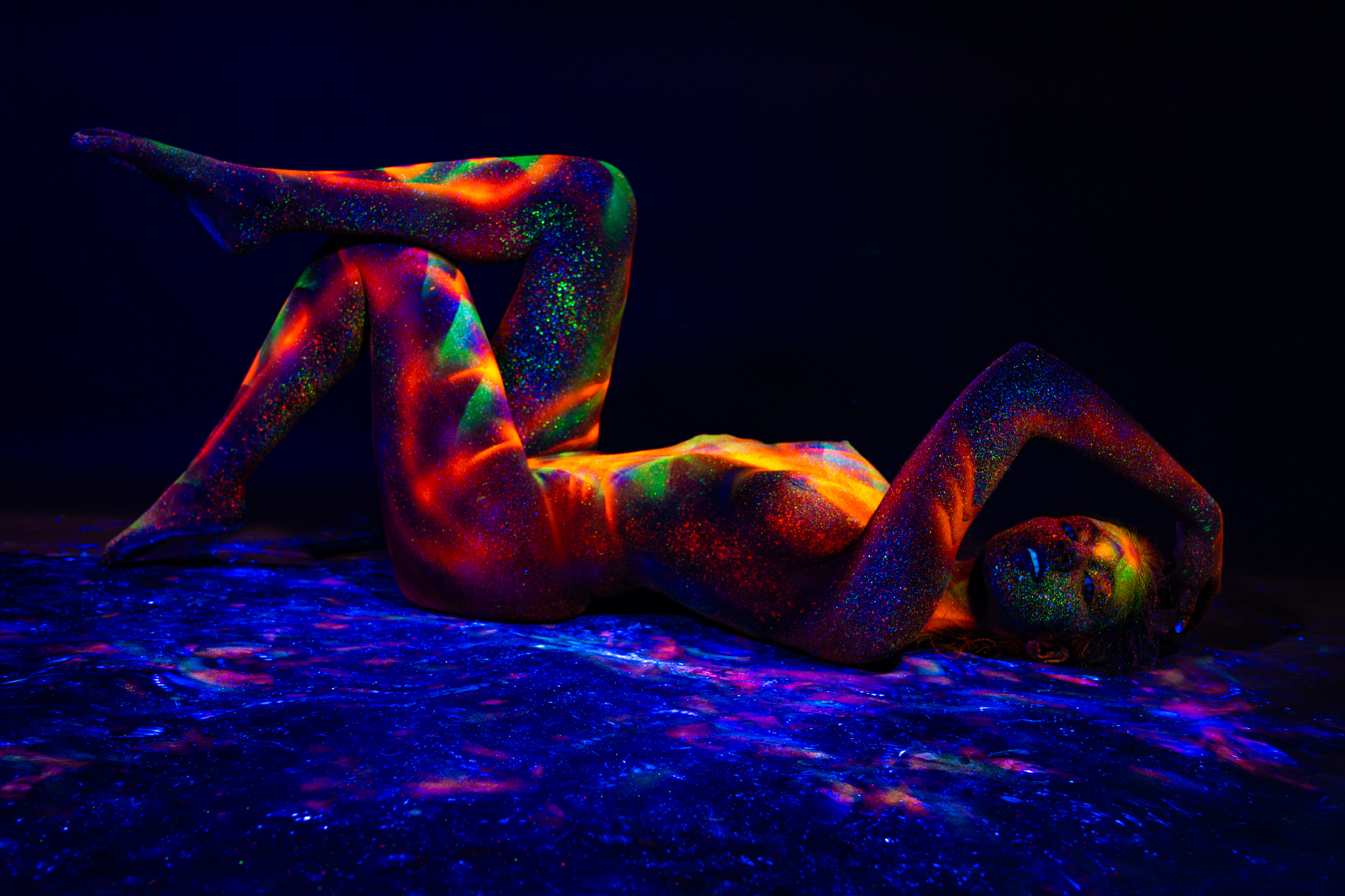 In this reclining pose, Sofia lies on her back with her knees bent and one leg lifted, head turned to the side on a reflective surface. A UV-reactive geometric design in orange, red, and green covers her body. Bodypaint by Elad Itzkin. Photographed under blacklight by Elad Itzkin Photography in his studio in Gesundbrunnen, Berlin.