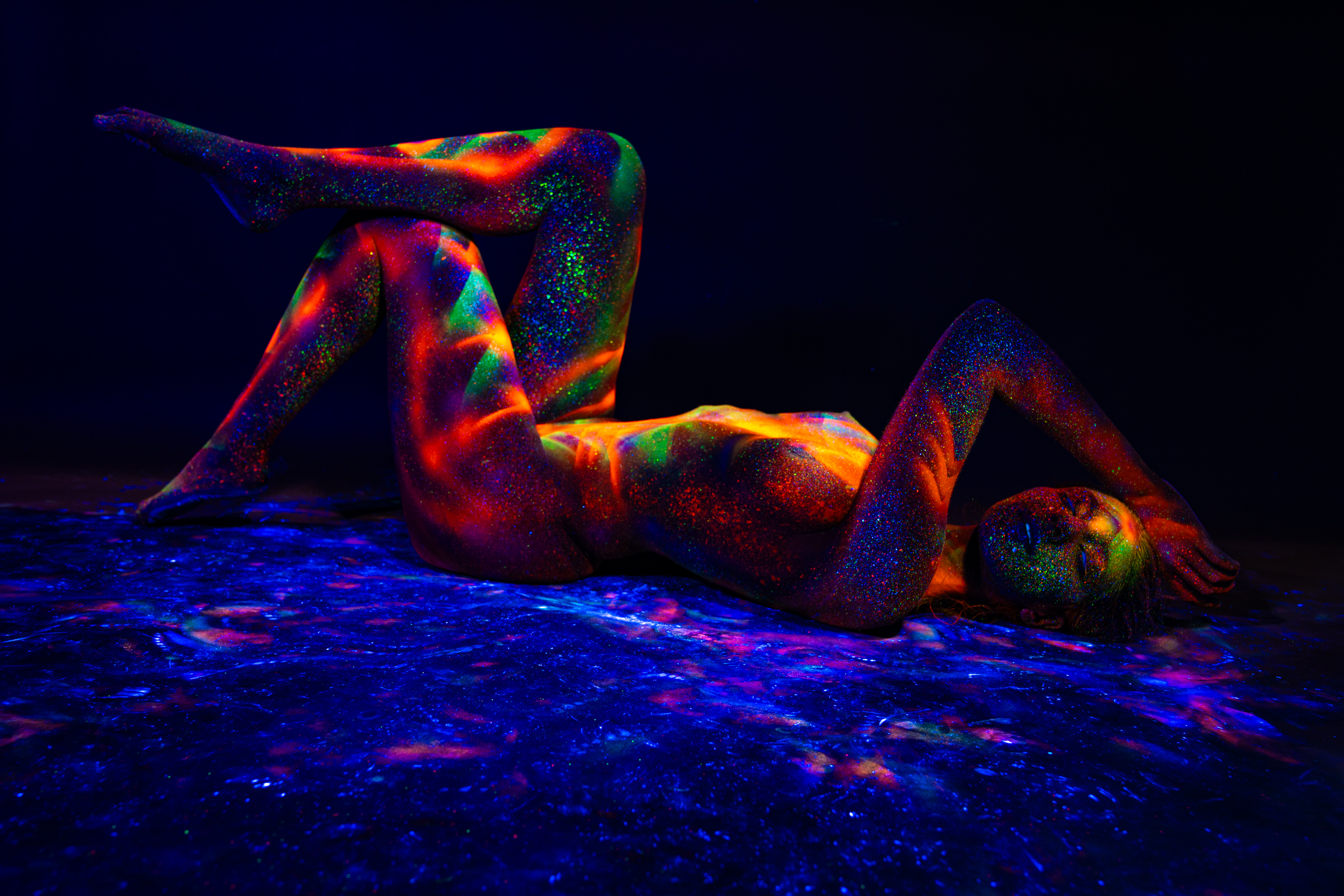 Sofia is photographed lying on her back with her knees bent and one leg raised, reclining on a reflective surface. Her torso and legs carry a UV-reactive geometric pattern in orange, red, and green tones. Bodypaint by Elad Itzkin. Photographed by Elad Itzkin Photography under UV light in his Gesundbrunnen, Berlin studio.