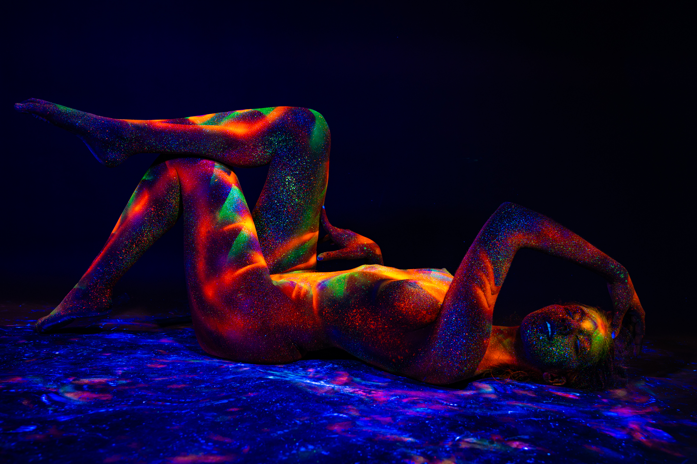 Sofia lies on her back with her knees bent and one leg extended upward, reclining on a reflective surface with her head turned to the side. Her body is covered in a UV-reactive geometric pattern in orange, red, and green. Bodypaint by Elad Itzkin. Photographed by Elad Itzkin Photography under UV light in his Gesundbrunnen, Berlin studio.