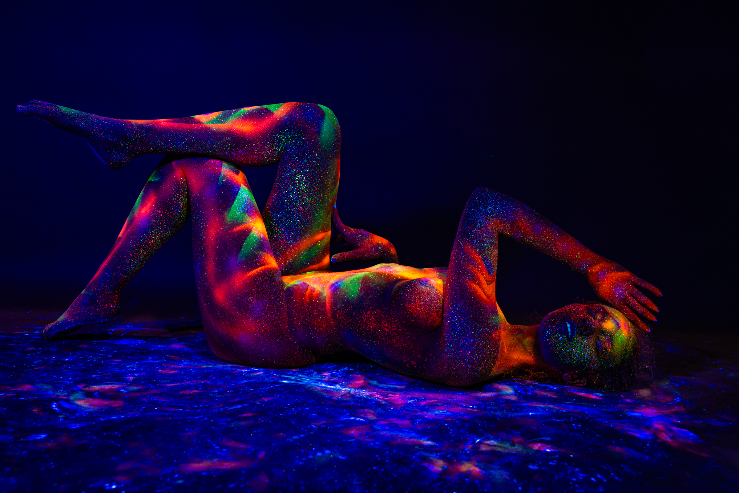 This reclining shot shows Sofia lying on her back with her knees bent, one leg raised, on a reflective surface. A UV-reactive geometric design in orange, red, and green tones covers her torso and legs. Bodypaint by Elad Itzkin. Photographed under blacklight by Elad Itzkin Photography in his studio in Gesundbrunnen, Berlin.