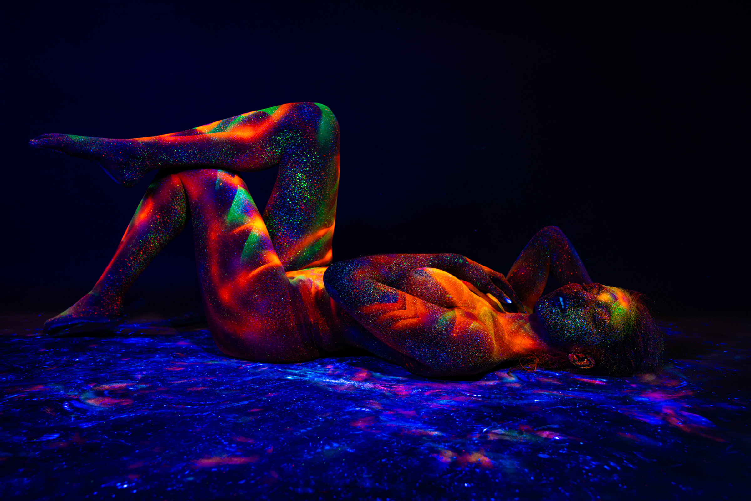 Sofia reclines on her back with one leg bent and raised, her upper body turned toward the camera. The design is a vivid geometric pattern of triangles and chevrons in orange, red, green and blue. Bodypaint by Elad Itzkin. Photographed by Elad Itzkin Photography under UV light in his Gesundbrunnen, Berlin studio.