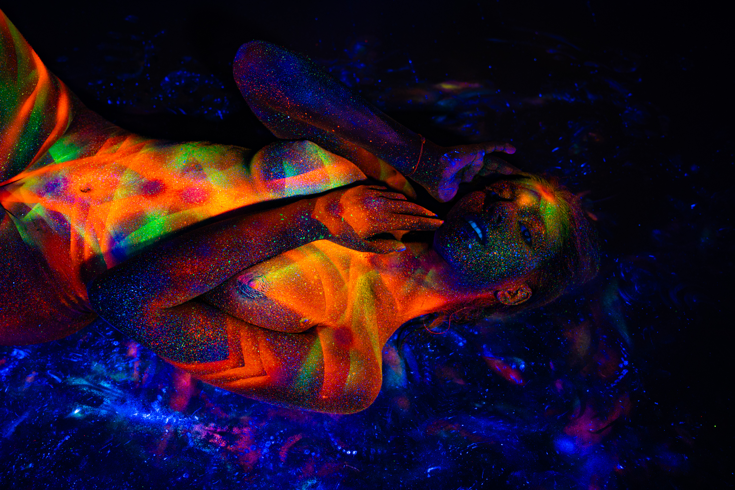 Sofia lies back in a reclining pose with one leg bent, her upper body and face angled toward the camera. Her skin is painted with a geometric chevron pattern in bright orange, red, green and blue. Bodypaint by Elad Itzkin. Photographed by Elad Itzkin Photography under UV light in his Gesundbrunnen, Berlin studio.
