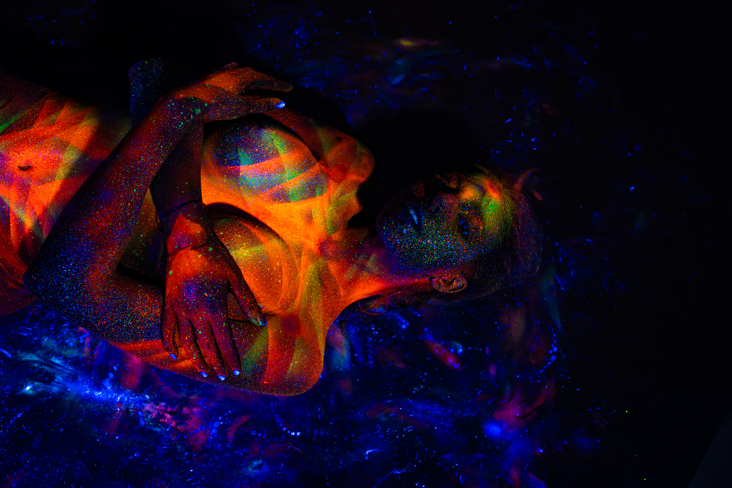 Sofia reclines on her back, framed closely on her upper body and face with one bent leg visible in the background. Her skin carries a geometric linework design in orange, red, green and blue. Bodypaint by Elad Itzkin. Photographed by Elad Itzkin Photography under UV light in his Gesundbrunnen, Berlin studio.