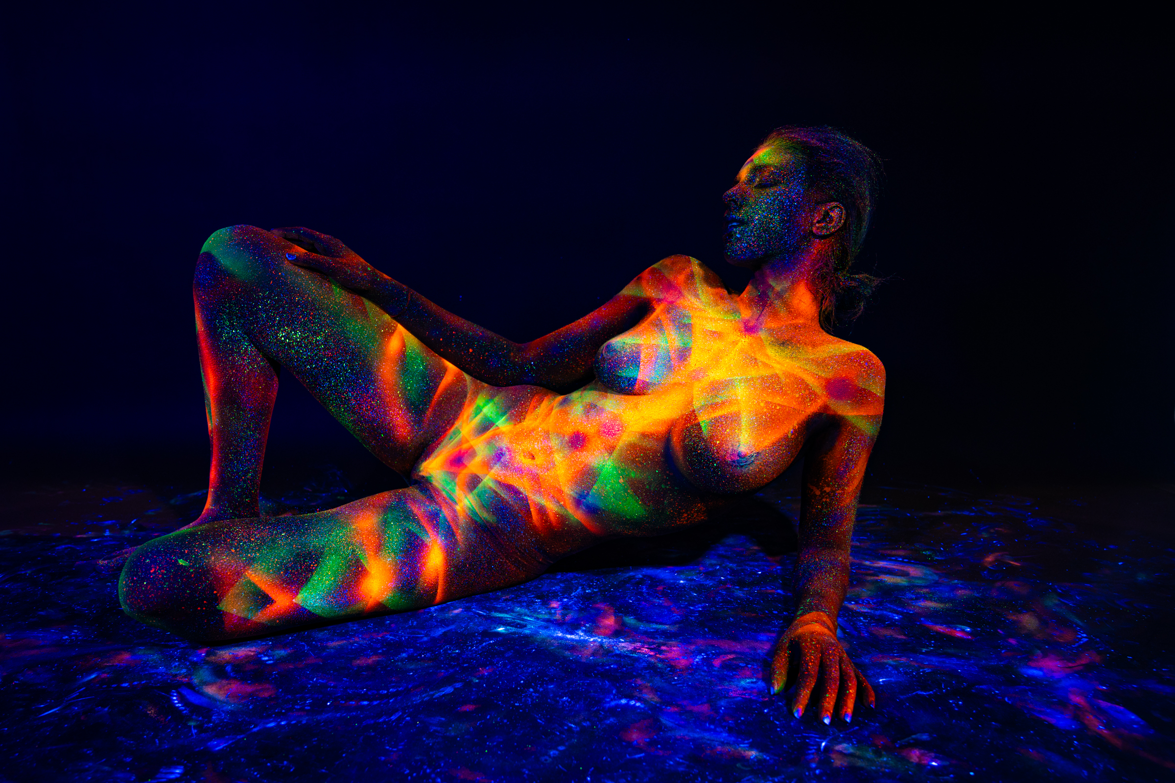 Sofia is in a semi-upright reclining pose with her torso facing forward and head tilted back. Her skin features a starburst-style geometric pattern in orange, green and blue tones. Bodypaint by Elad Itzkin. Photographed by Elad Itzkin Photography under UV light in his Gesundbrunnen, Berlin studio.