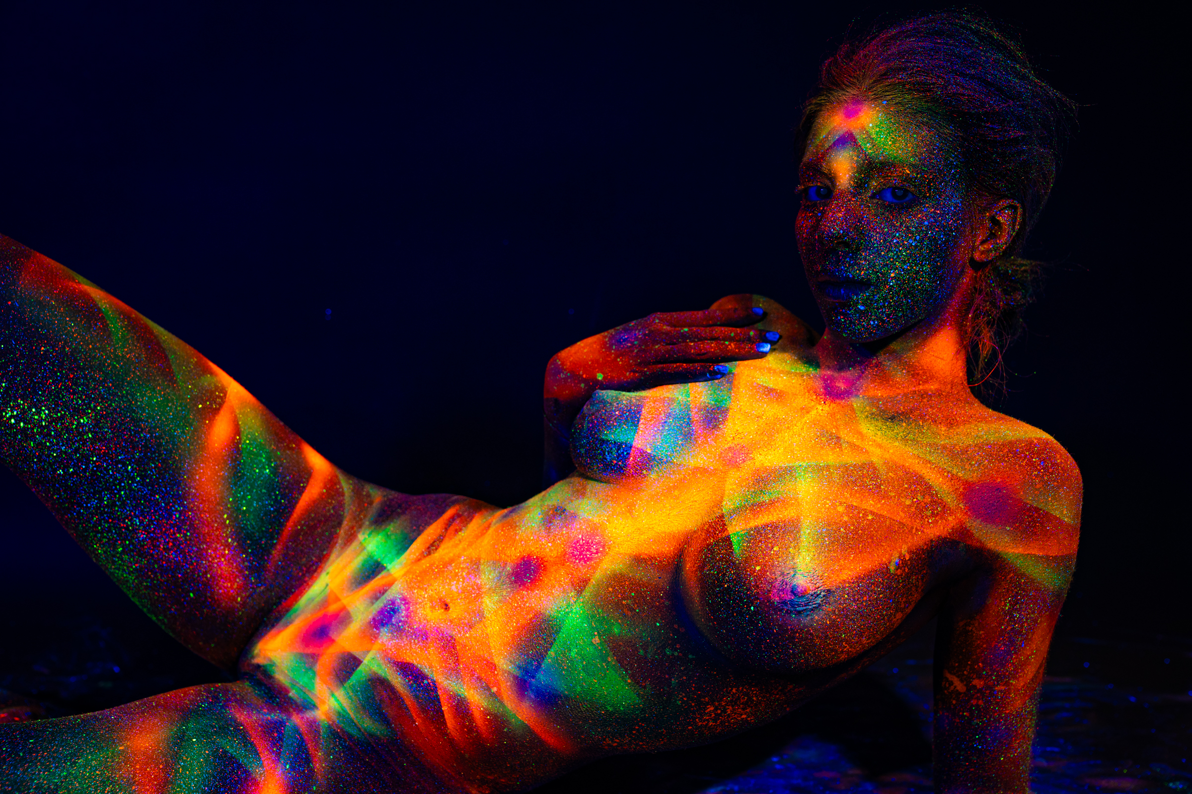 Sofia reclines in a semi-upright position, her torso turned toward the camera and head tilted to the side. A geometric pattern of interlocking lines in orange, green and blue covers her chest. Bodypaint by Elad Itzkin. Photographed by Elad Itzkin Photography under UV light in his Gesundbrunnen, Berlin studio.