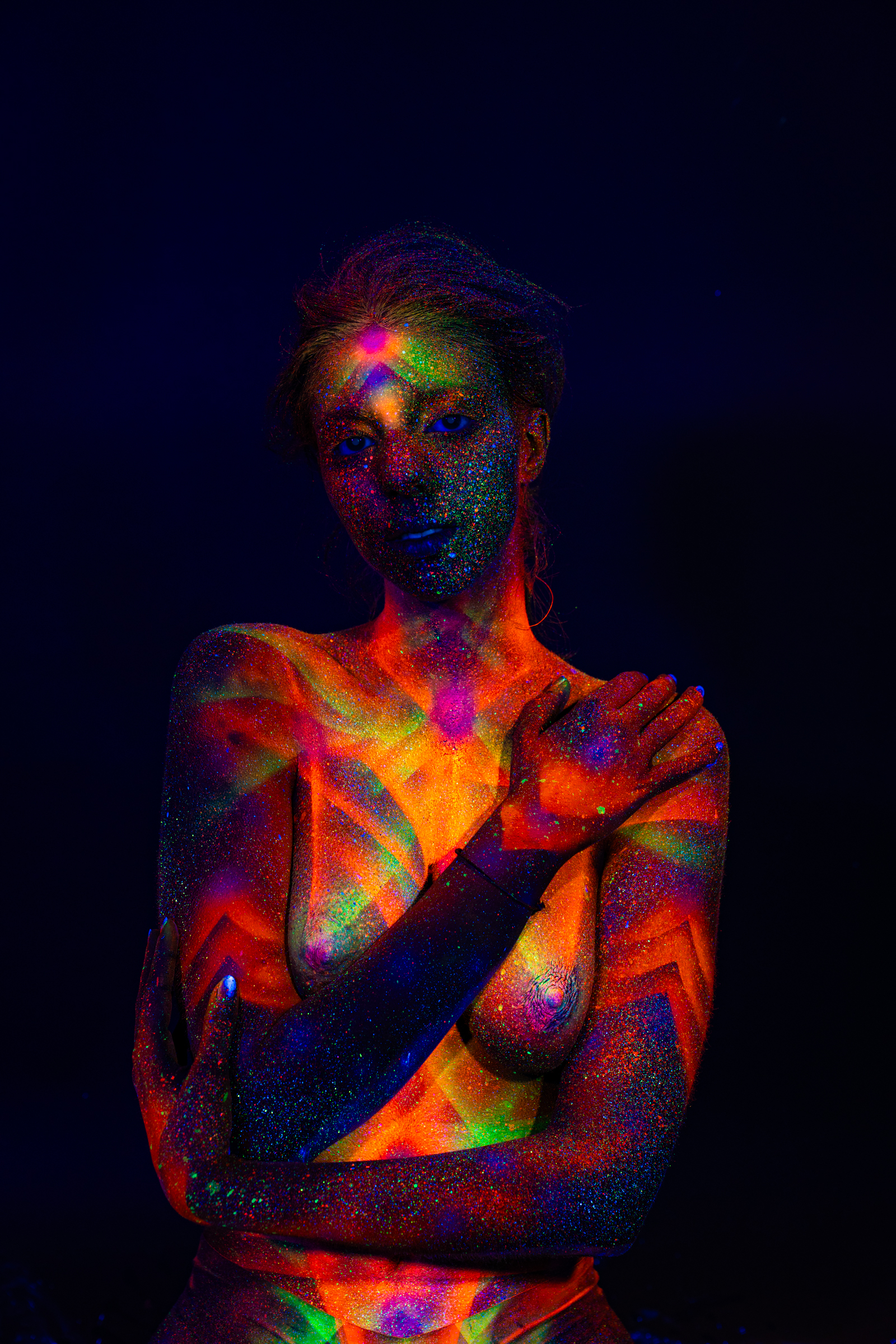 Sofia is captured in a three-quarter portrait pose, her upper body angled slightly to the side. Her chest and shoulders are covered in a geometric linework pattern in orange, green and blue. Bodypaint by Elad Itzkin. Photographed by Elad Itzkin Photography under UV light in his Gesundbrunnen, Berlin studio.