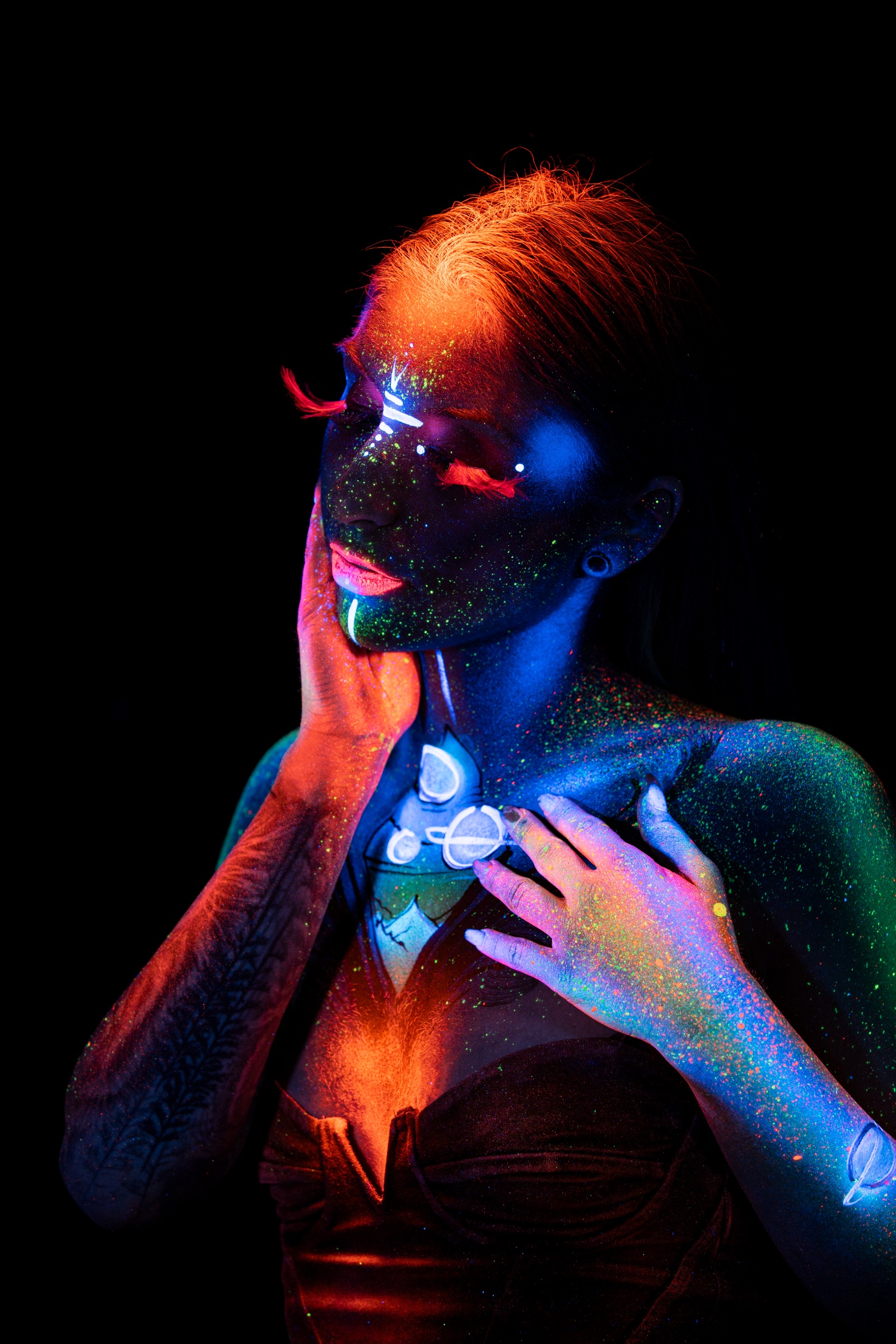 Sori stands with her head turned to the side in a three-quarter pose. A cosmic design in blue and orange featuring planet and ring motifs covers her chest, paired with dark UV face paint. Bodypaint by Claudia Kraemer. Photographed by Elad Itzkin Photography under blacklight in his Gesundbrunnen, Berlin studio.